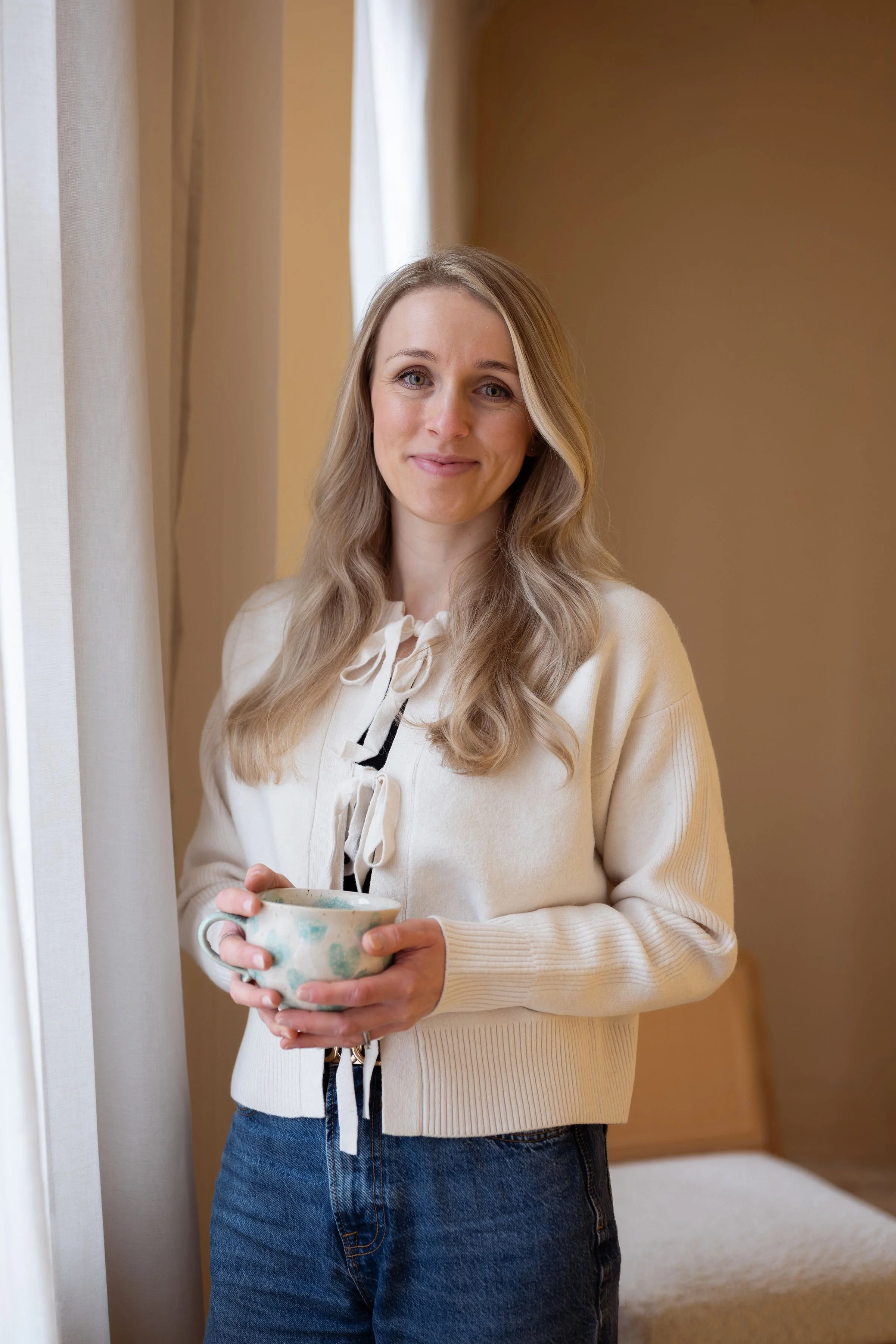 A woman with long wavy blonde hair holding a ceramic mug, standing indoors near a window with curtains, wearing a cream-colored sweater and jeans, smiling at the camera.