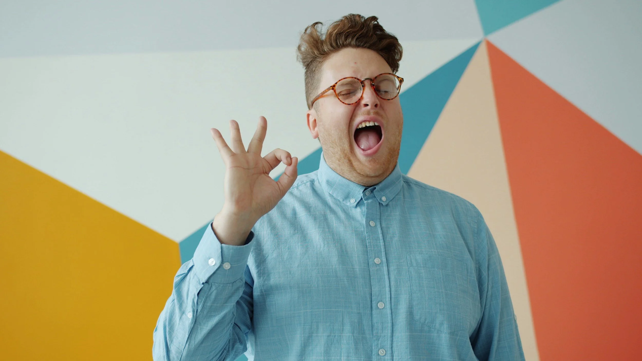 A man with curly hair, glasses, and a light blue button-up shirt making an OK gesture with his hand while yawning in front of a colorful, geometric background.