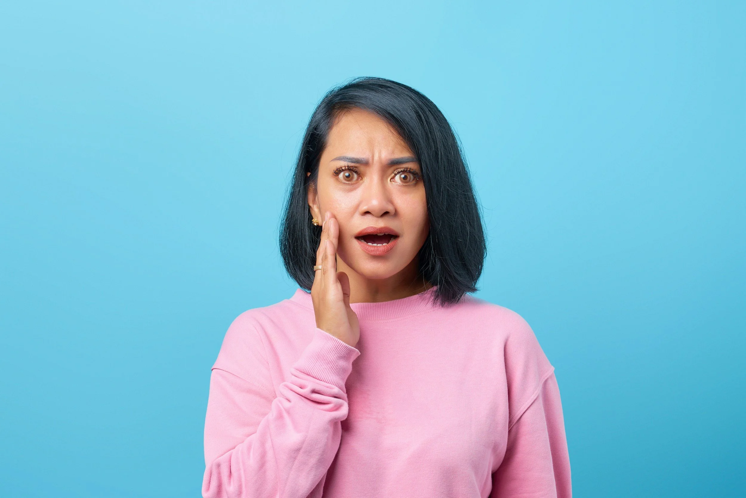 A woman with black hair and light brown skin, wearing a pink sweater, holding her cheek with a surprised or worried expression on her face, standing against a blue background.