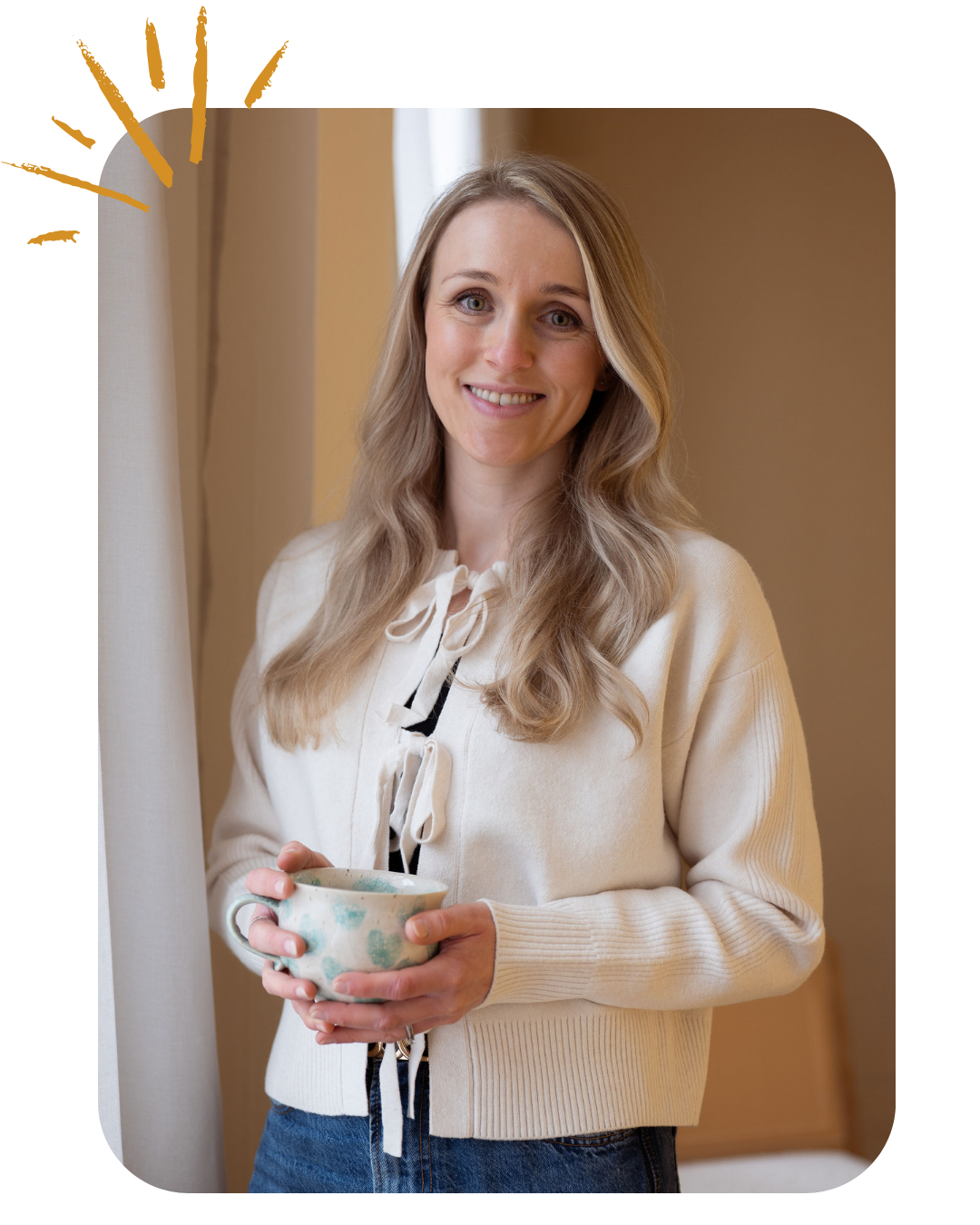 A young woman with long blonde hair, smiling, holding a mug, standing near a window with curtains in a warmly lit room.