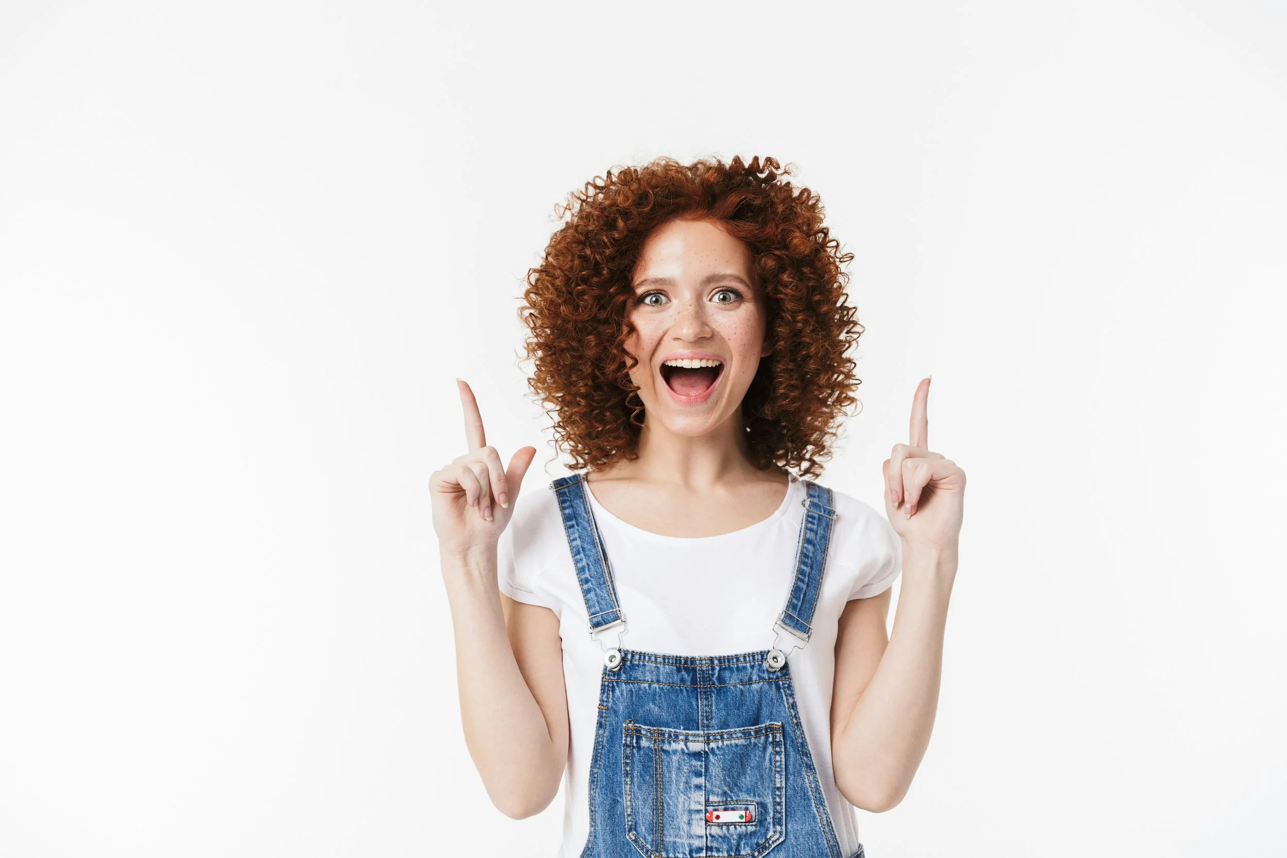 A young woman with curly red hair, smiling and pointing upward with both index fingers, wearing a white t-shirt and blue denim overalls, standing against a plain white background.