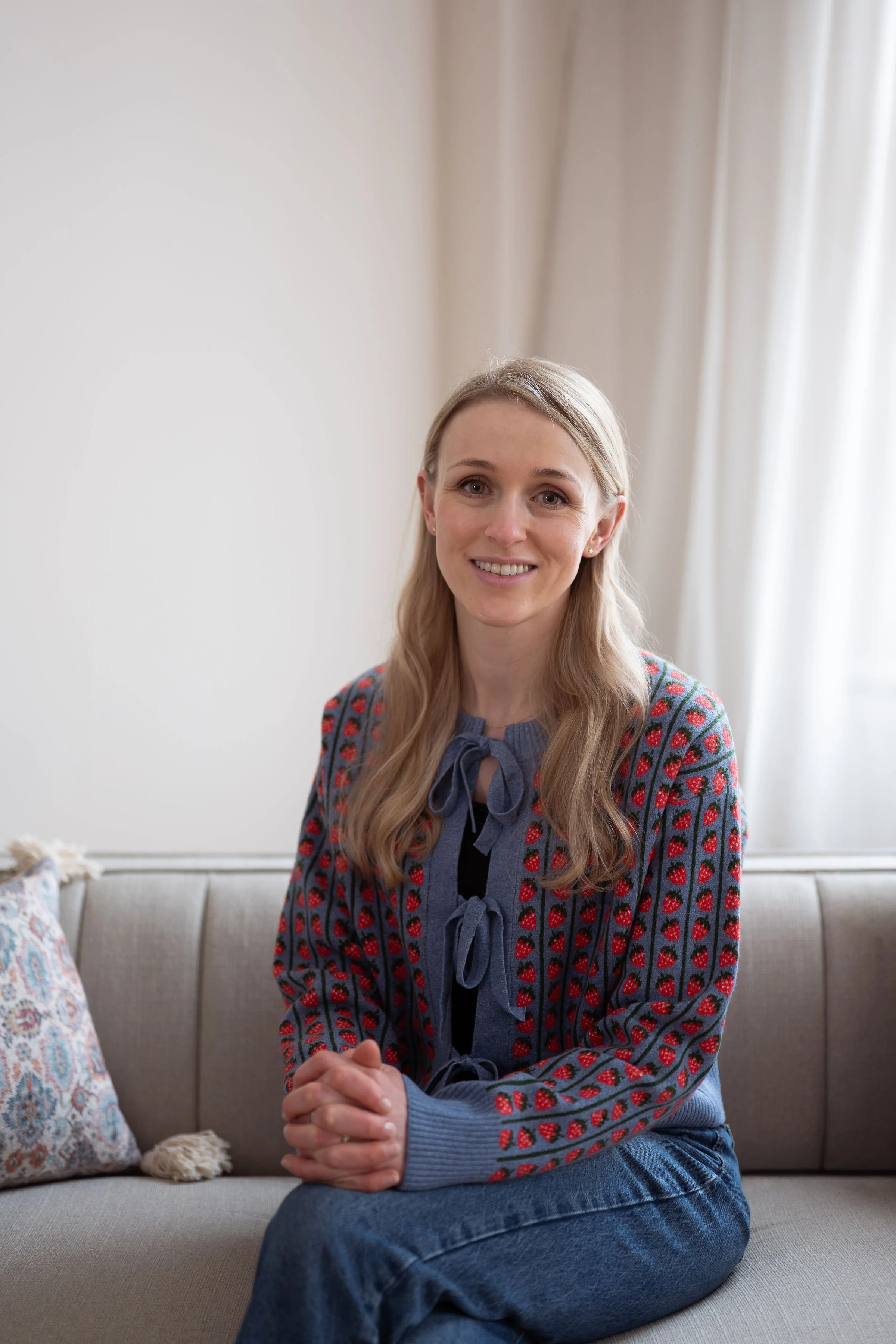 A woman with long blonde hair sitting on a beige sofa, wearing a blue sweater with a bear pattern and a dark blue bow. She is smiling and clasping her hands.