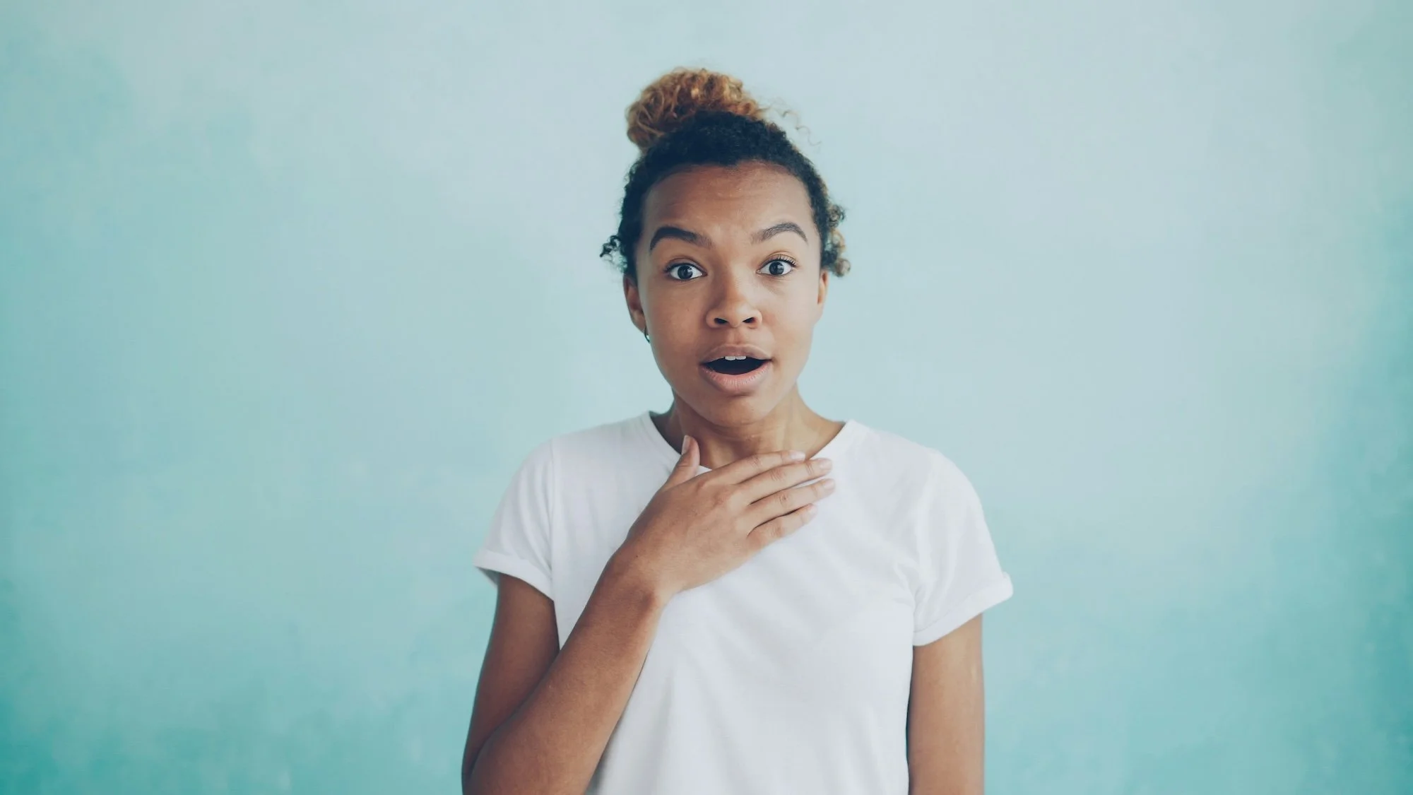 A young woman with curly hair in a bun, wearing a white t-shirt, is touching her throat with her right hand and has a surprised expression.