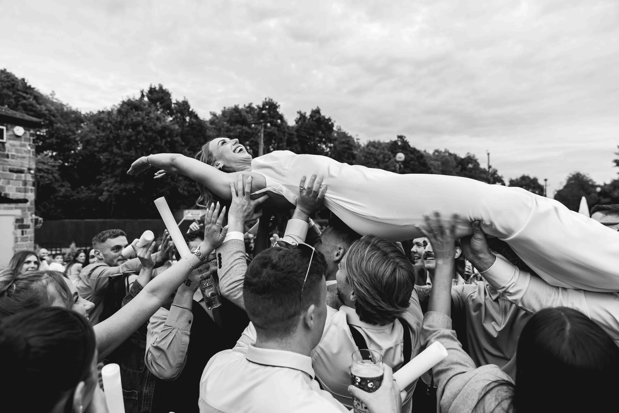 People outdoors celebrating as a woman in a white dress is lifted above their heads during a special event or wedding, with trees and a cloudy sky in the background.