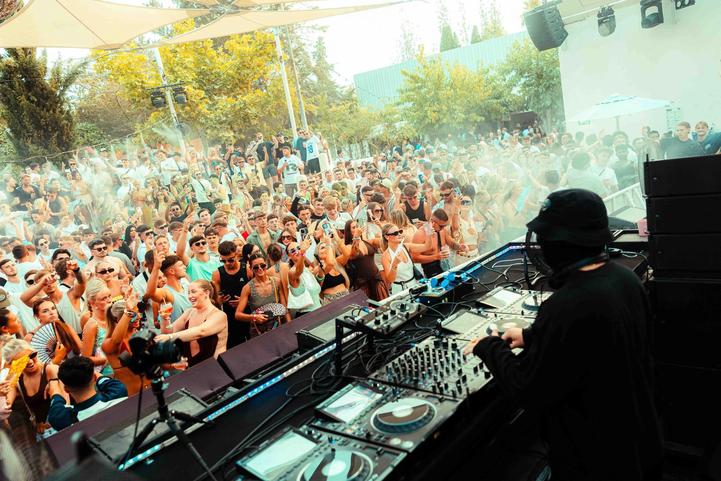 A large crowd of people attending an outdoor music festival or event, seen from behind the DJ booth, with DJ equipment in the foreground. The audience appears to be enjoying the event, with many people taking photos or videos with their phones, and s
