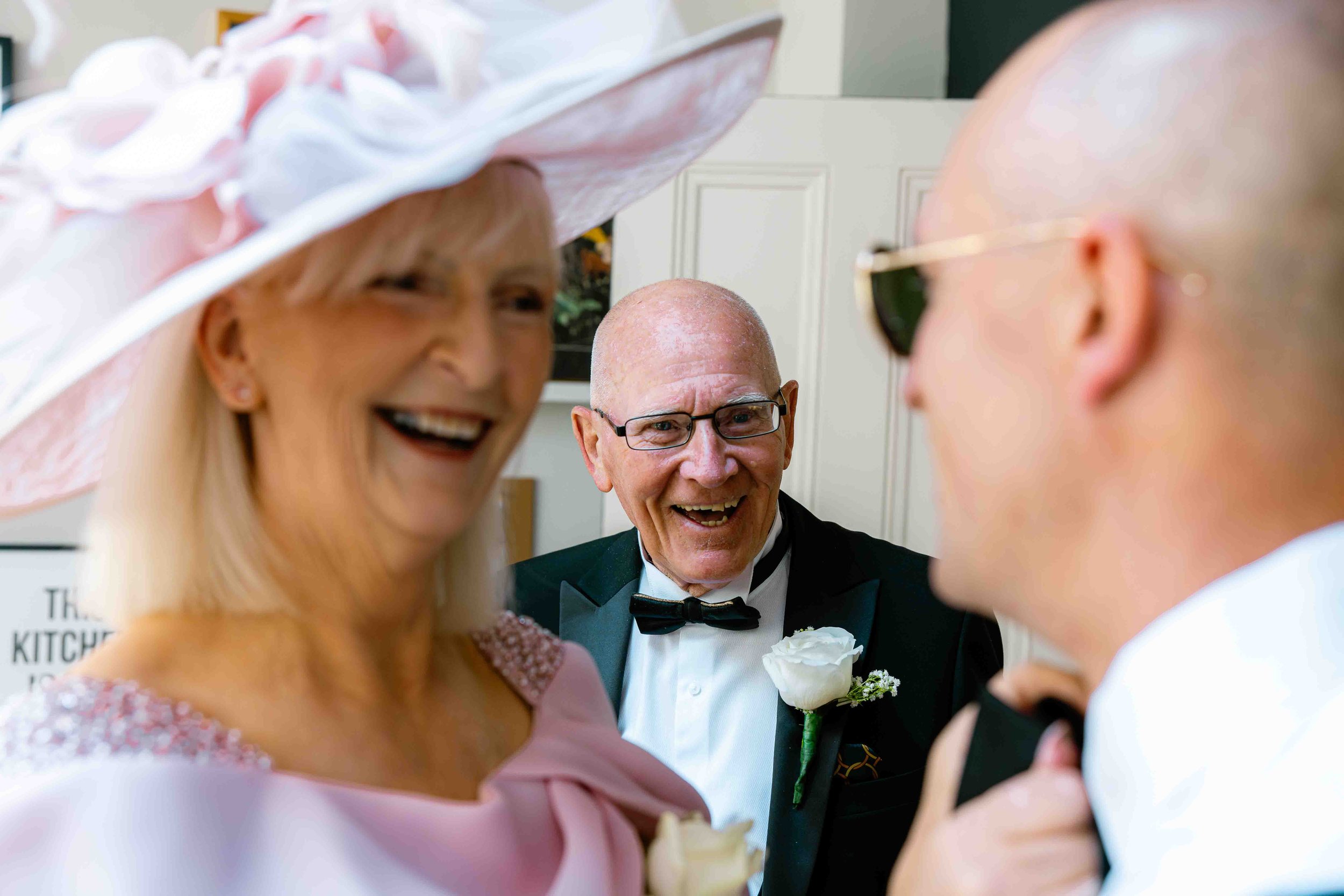 A wedding scene with a bride, groom, and a smiling older man in tuxedo, in an indoor setting.