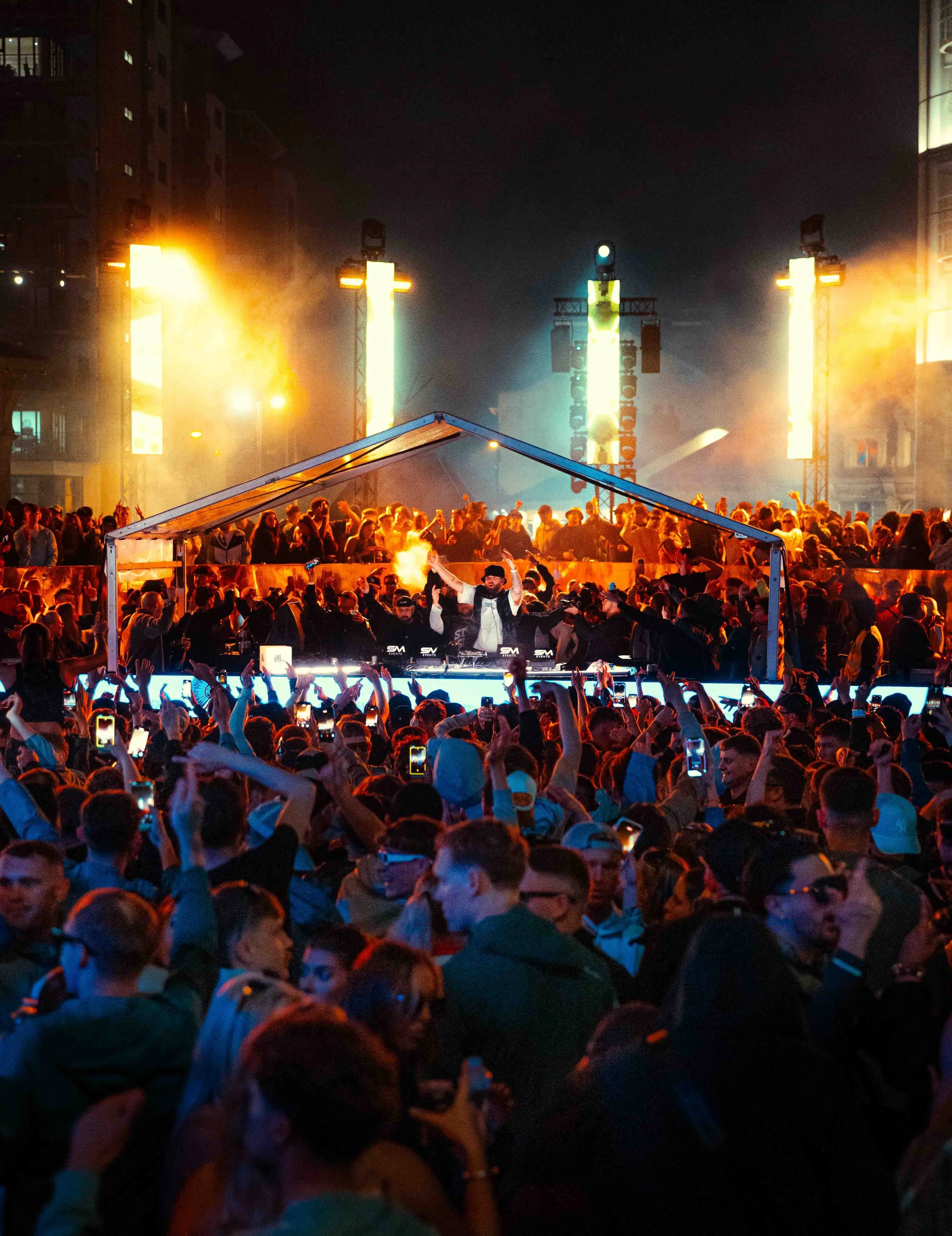 Gaskin plays to a crowded outdoor music concert at night with a DJ in the foreground, bright stage lights, large screens, and a diverse audience raising their hands and phones.