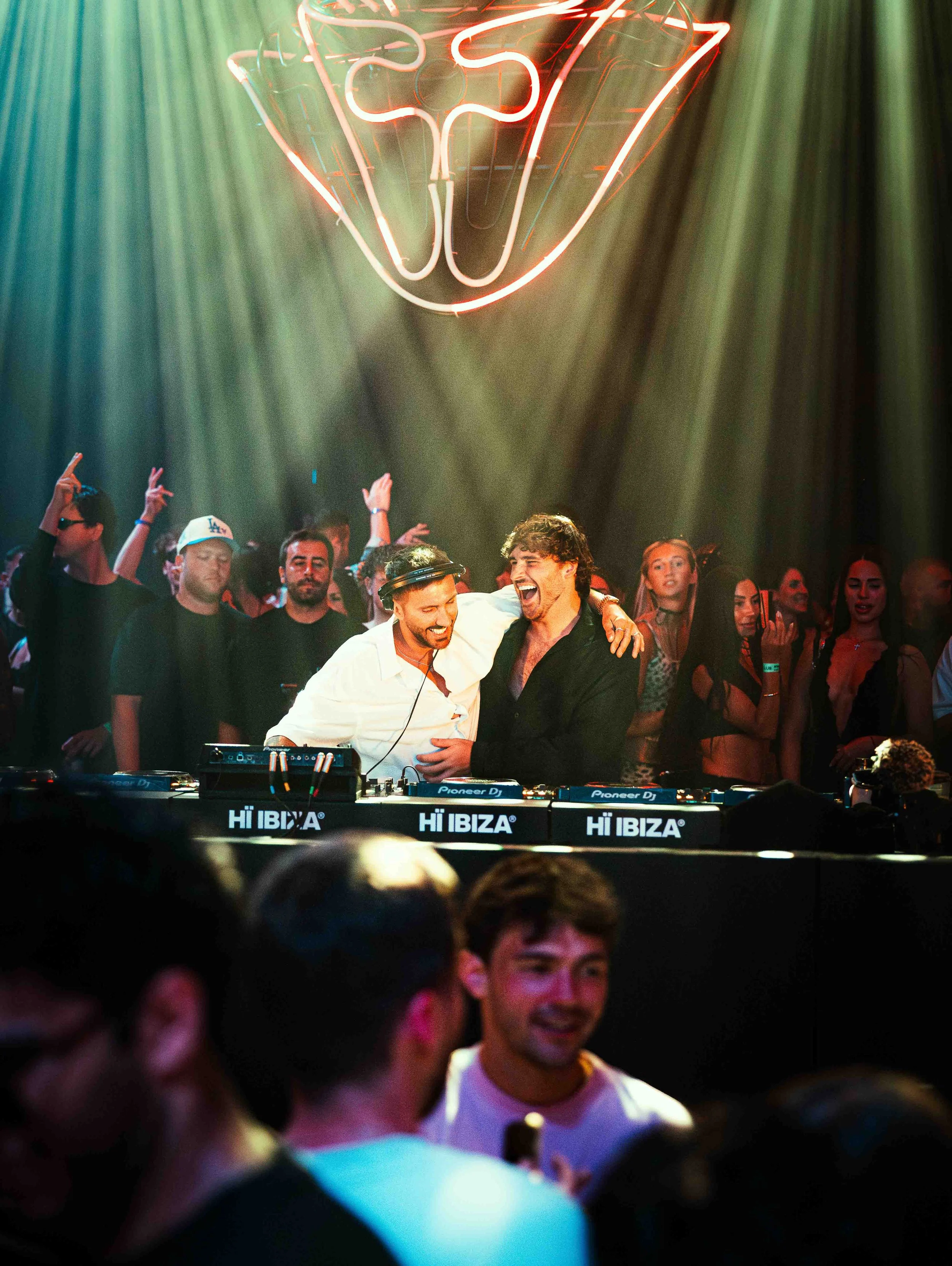 A group of people dancing and smiling at a nightclub or party, with a neon sign hanging above them and a DJ booth in the foreground.