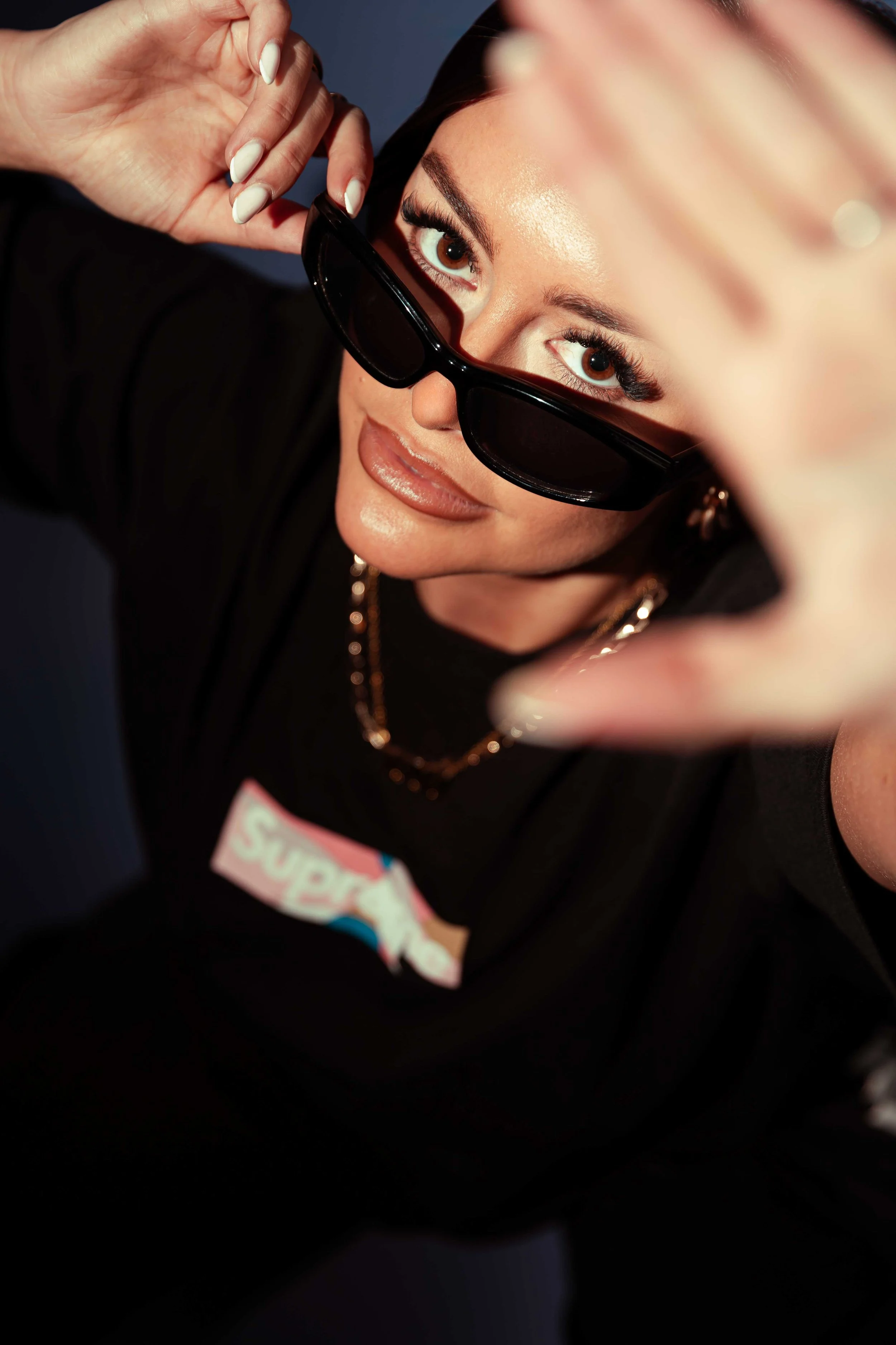Close-up of a woman with light skin and brown eyes, wearing black sunglasses slightly lowered on her nose, looking up at the camera. She has dark hair and makeup with glossy lips. The background is dark and blurred.