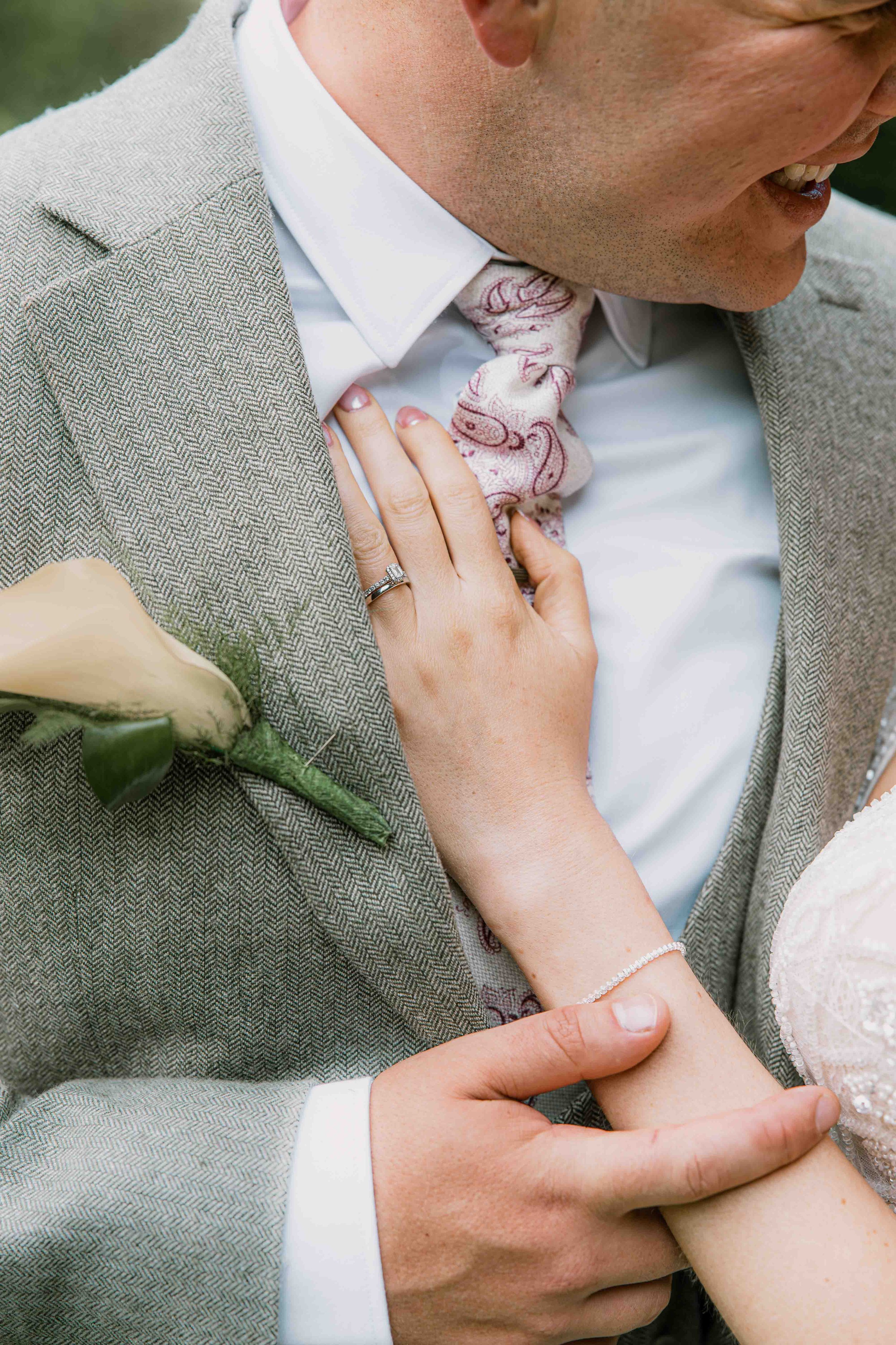 Close-up of a wedding scene showing a man and woman holding hands, the man's hand on the woman's arm, with the woman wearing a wedding ring and a delicate bracelet, and a white rose with green leaves pressed against the man's gray herringbone suit jacket, smiling and touching each other affectionately.