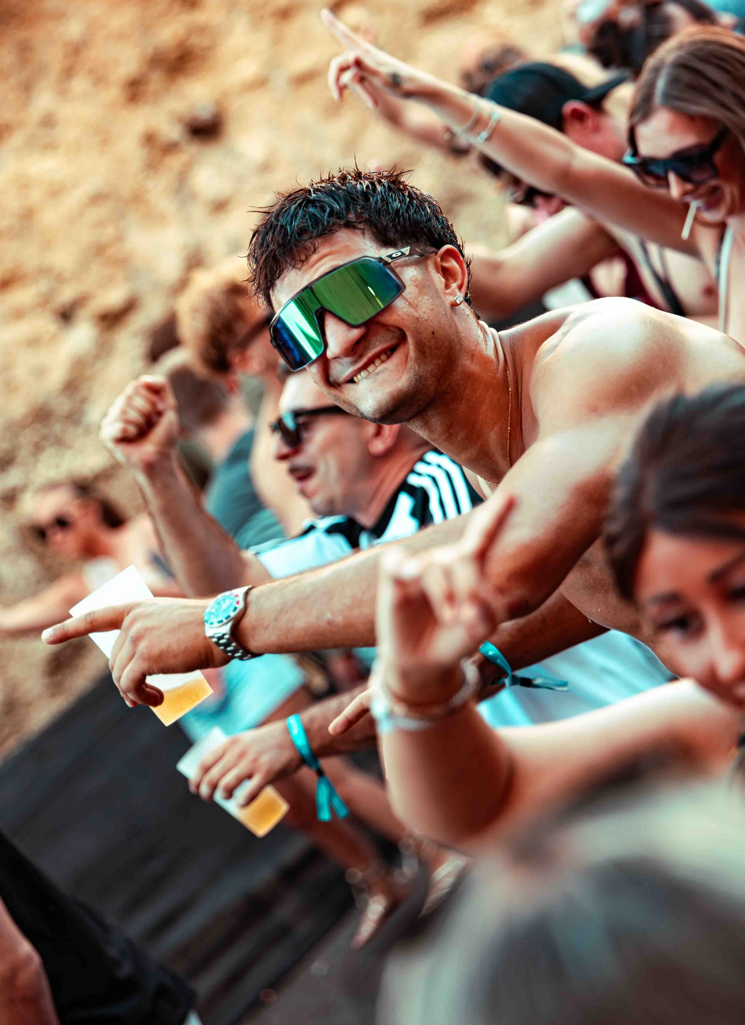 People sitting in a row at an outdoor event, some wearing sunglasses and wristbands, smiling and holding drinks, with a sandy or gravel background.