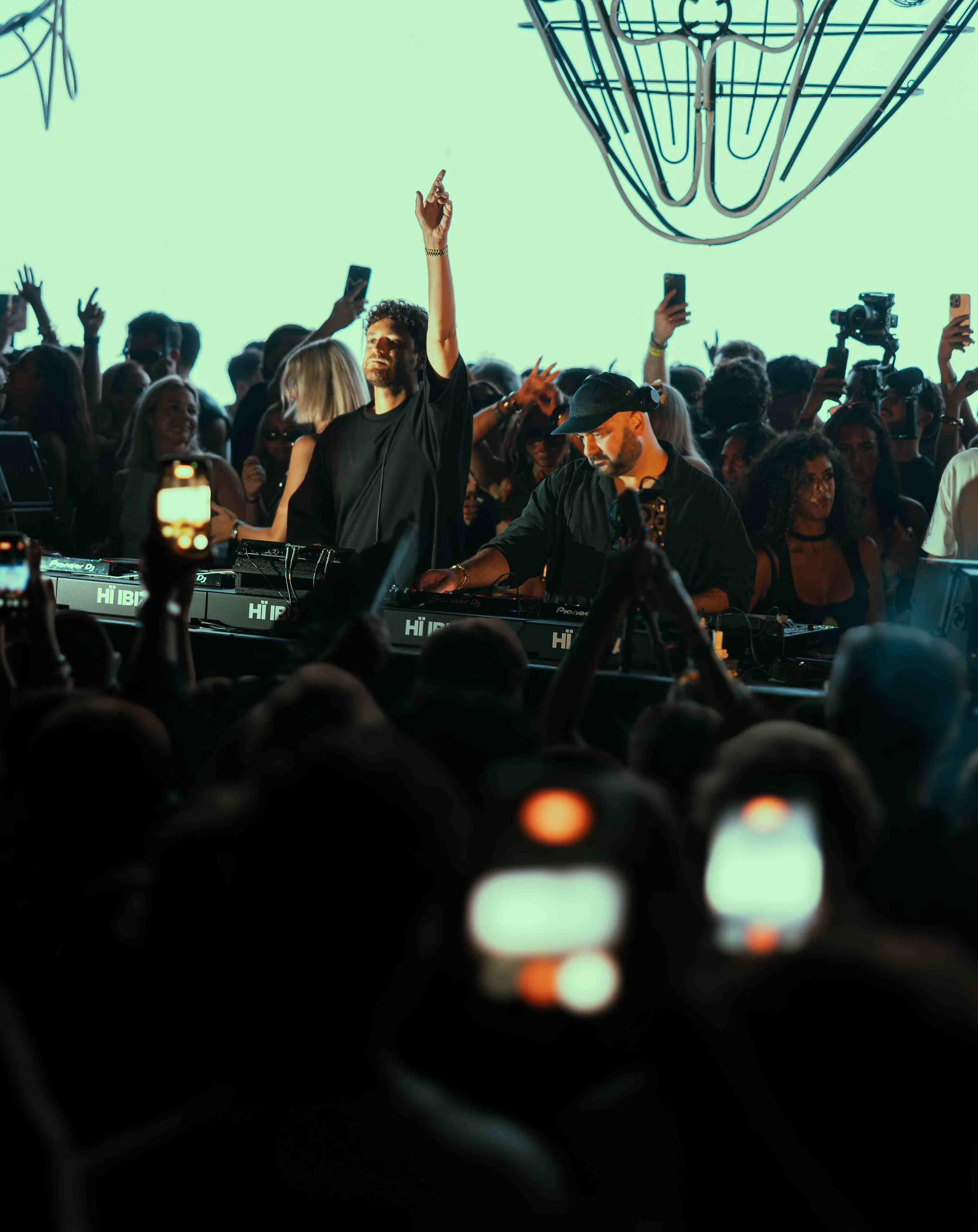 A DJ performing at a crowded event with people taking photos and videos with their smartphones, and a large wall light installation in the background.