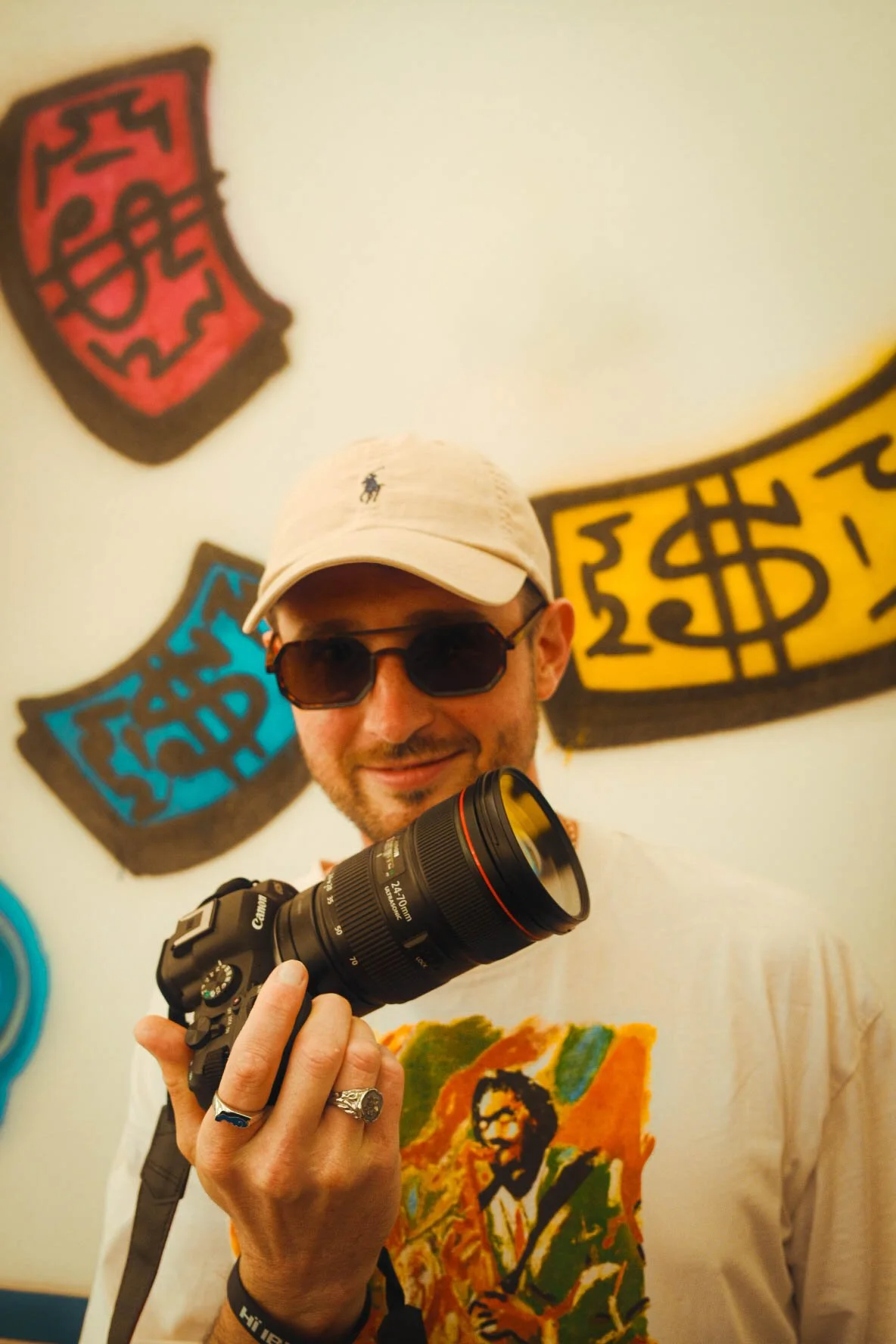A man wearing sunglasses and a beige baseball cap holding a camera with a large lens, smiling indoors in front of wall art featuring colorful, stylized dollar signs and a portrait of a woman.