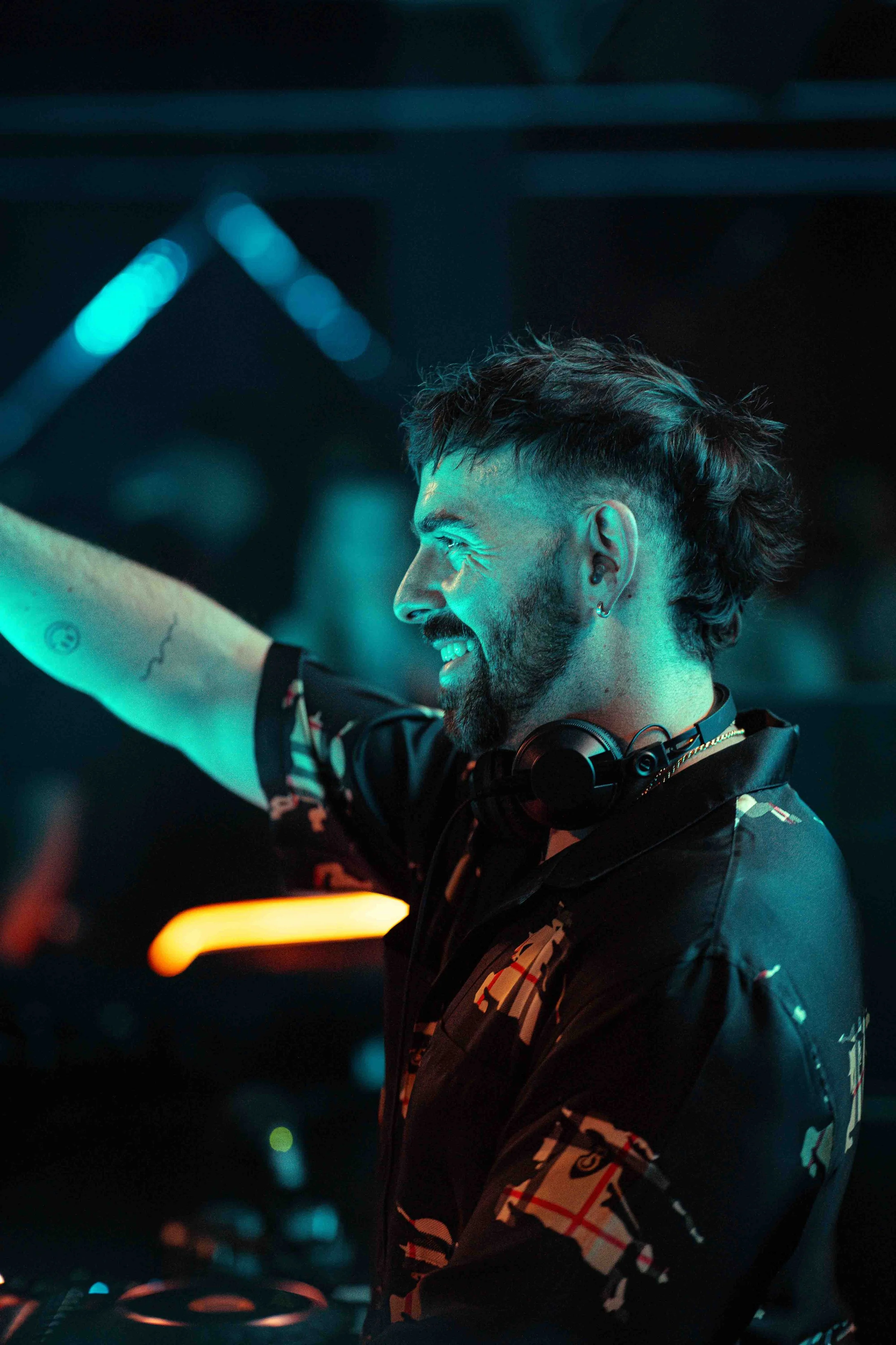 A young man with a beard and styled hair smiling and dancing at night, wearing headphones around his neck in a dimly lit setting with colorful lights.