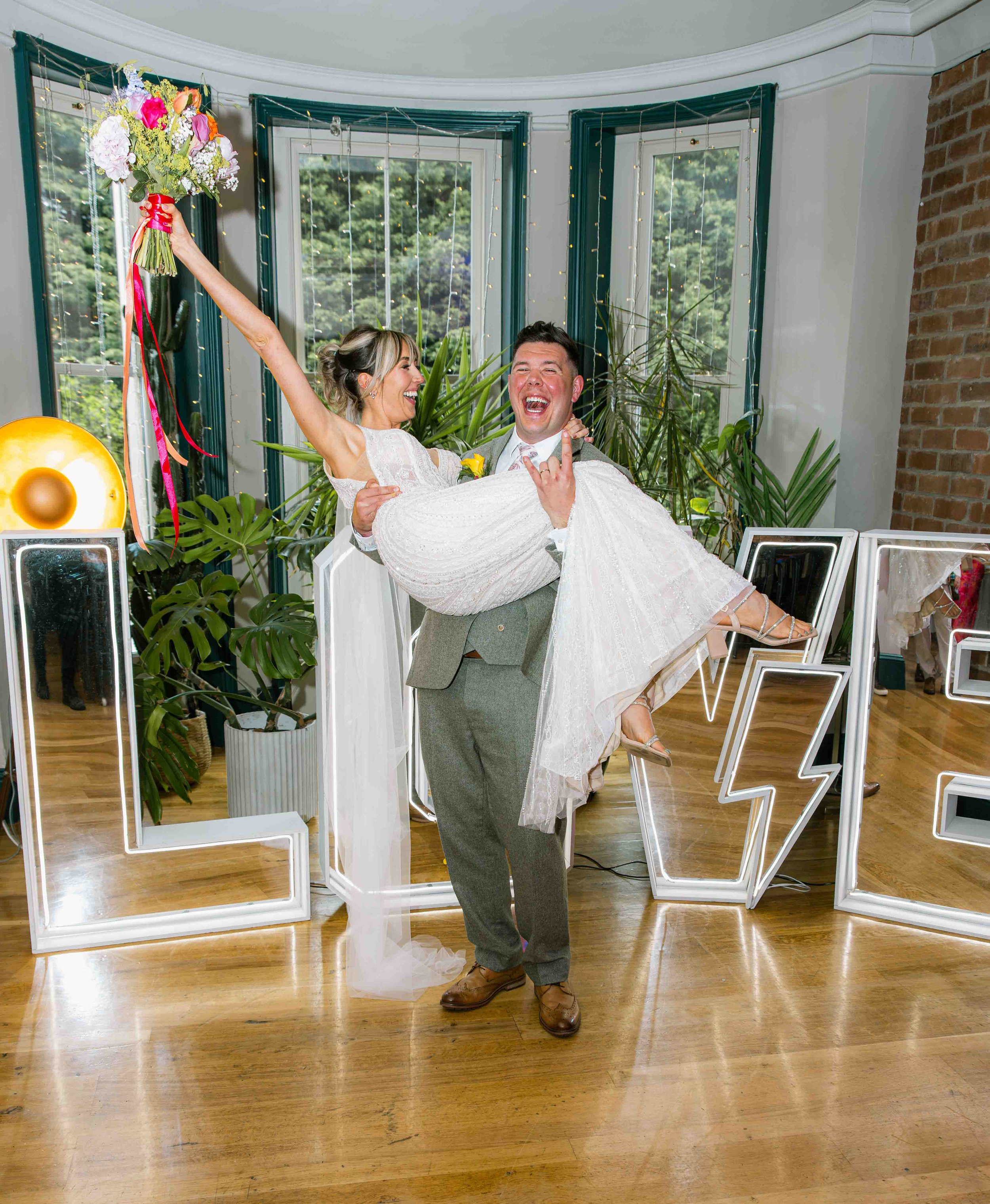 A joyful bride in a white wedding dress holding a bouquet of flowers, being swung around by a groom in a gray suit with a white shirt and tie, in a decorated indoor space with large windows, green plants, and decorative lighting.