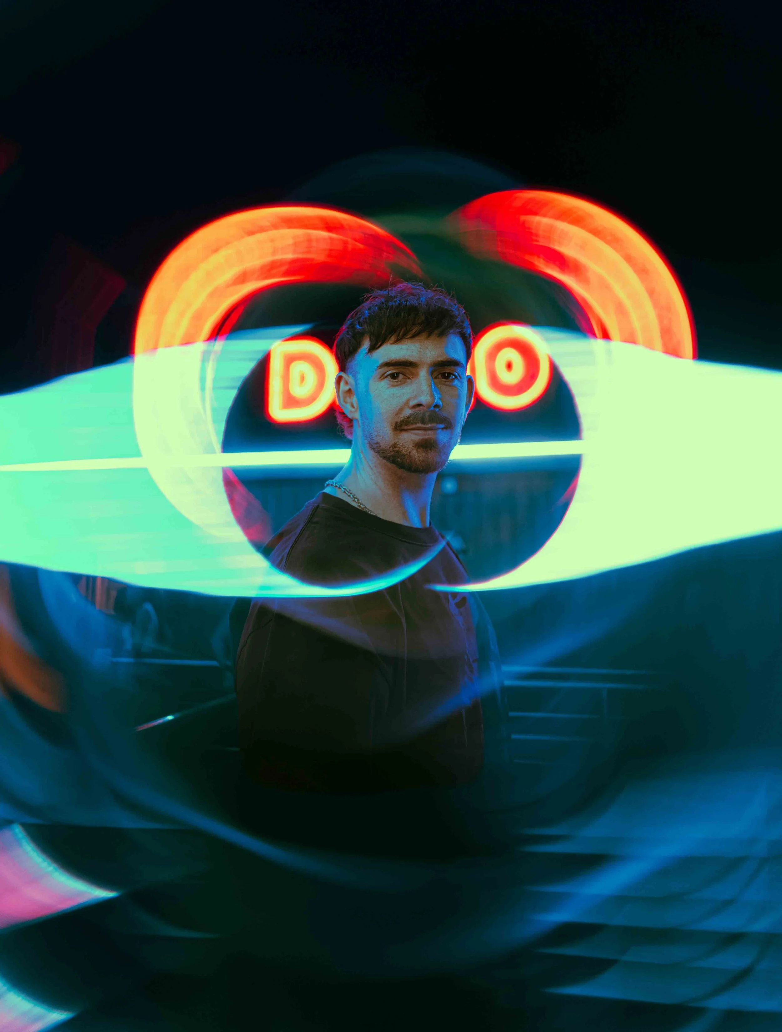 Portrait of a young man with dark hair and facial hair, illuminated by colorful neon lights, with blurred vibrant light trails in the background.