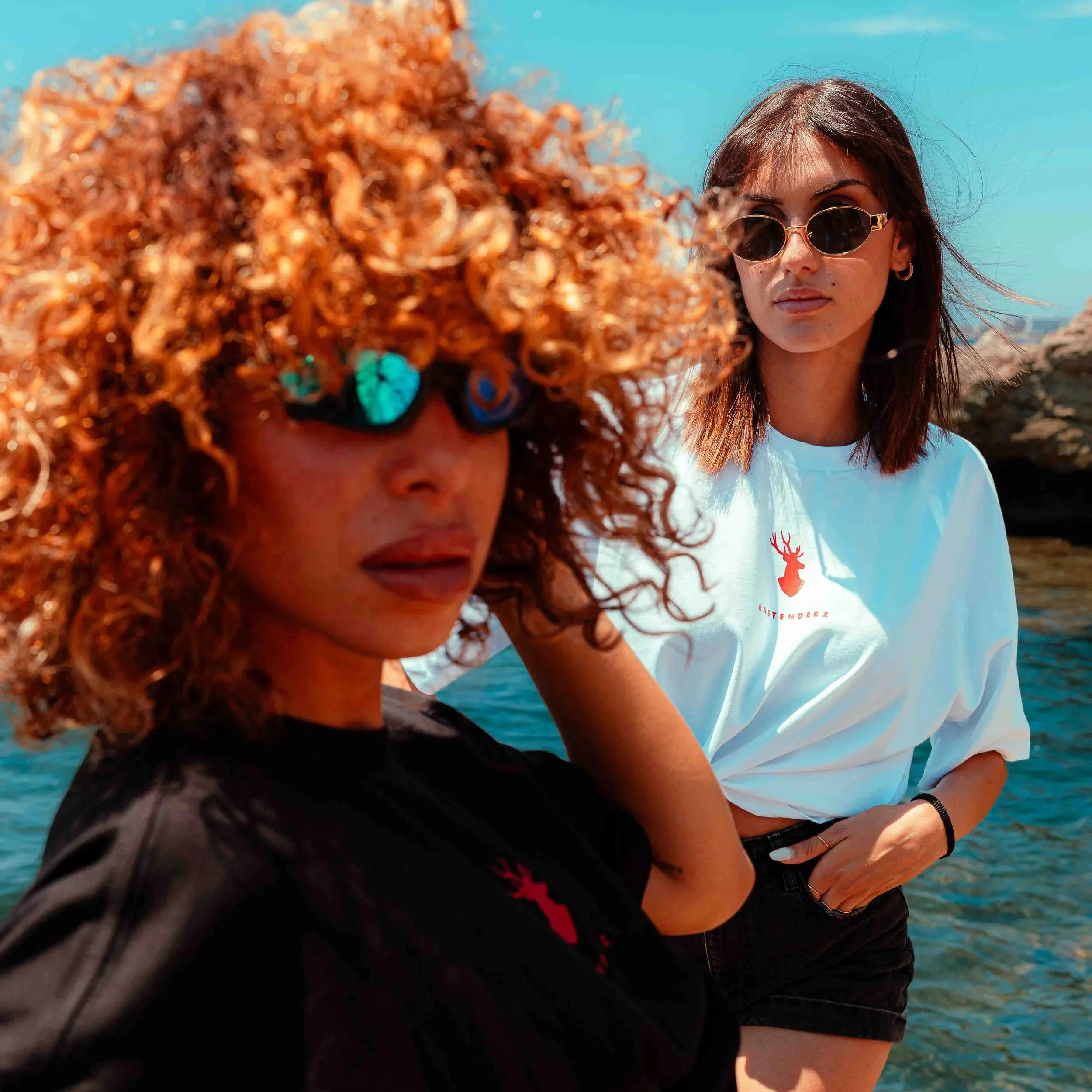 A woman with curly red hair and dark skin, wearing sunglasses, is partly visible in the foreground. In the background, a woman with light skin and straight brown hair, wearing sunglasses and a white t-shirt with a red logo, stands outdoors under a cl