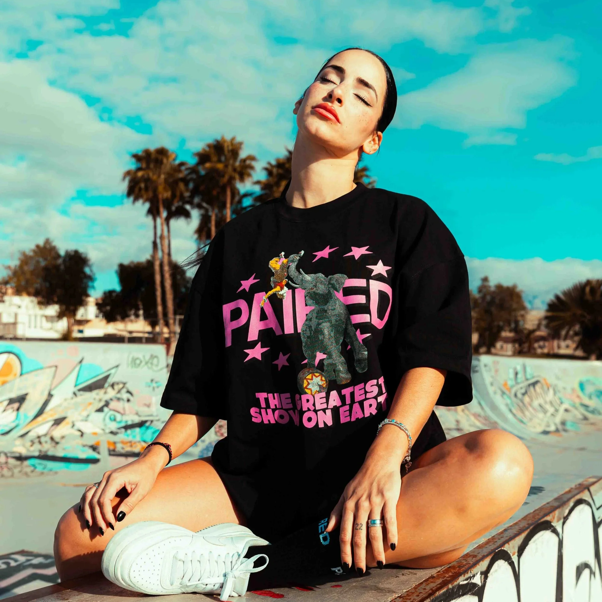 A young woman with dark hair, freckles, wearing a black T-shirt with pink stars and text, white sneakers, earrings, and rings, sitting on a graffiti-covered rail with palm trees and a blue sky in the background, eyes closed and face relaxed.