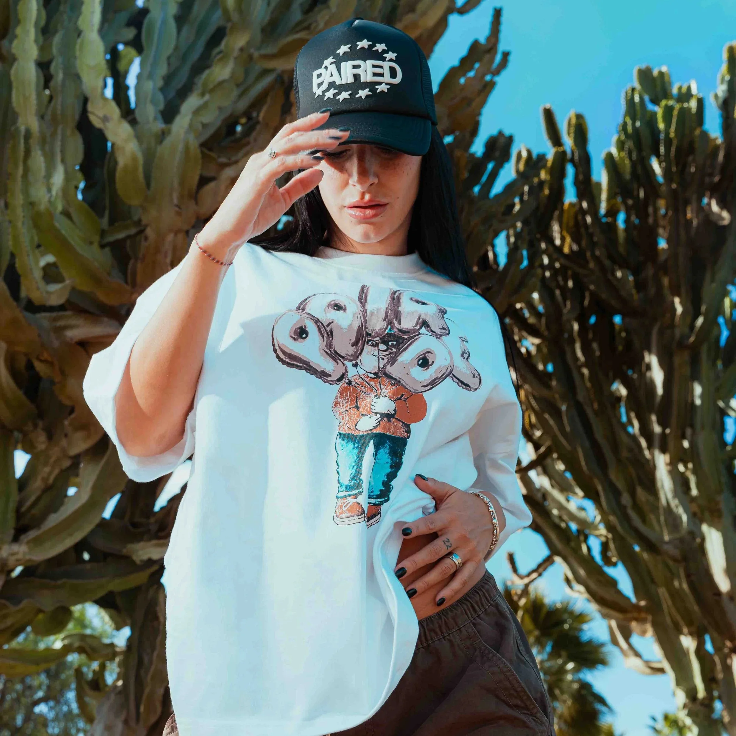 A woman wearing a black cap with the word 'PAIRED' and stars above it features her standing outdoors against a background of tall, green succulents and bright blue sky, with her hand on her waist.