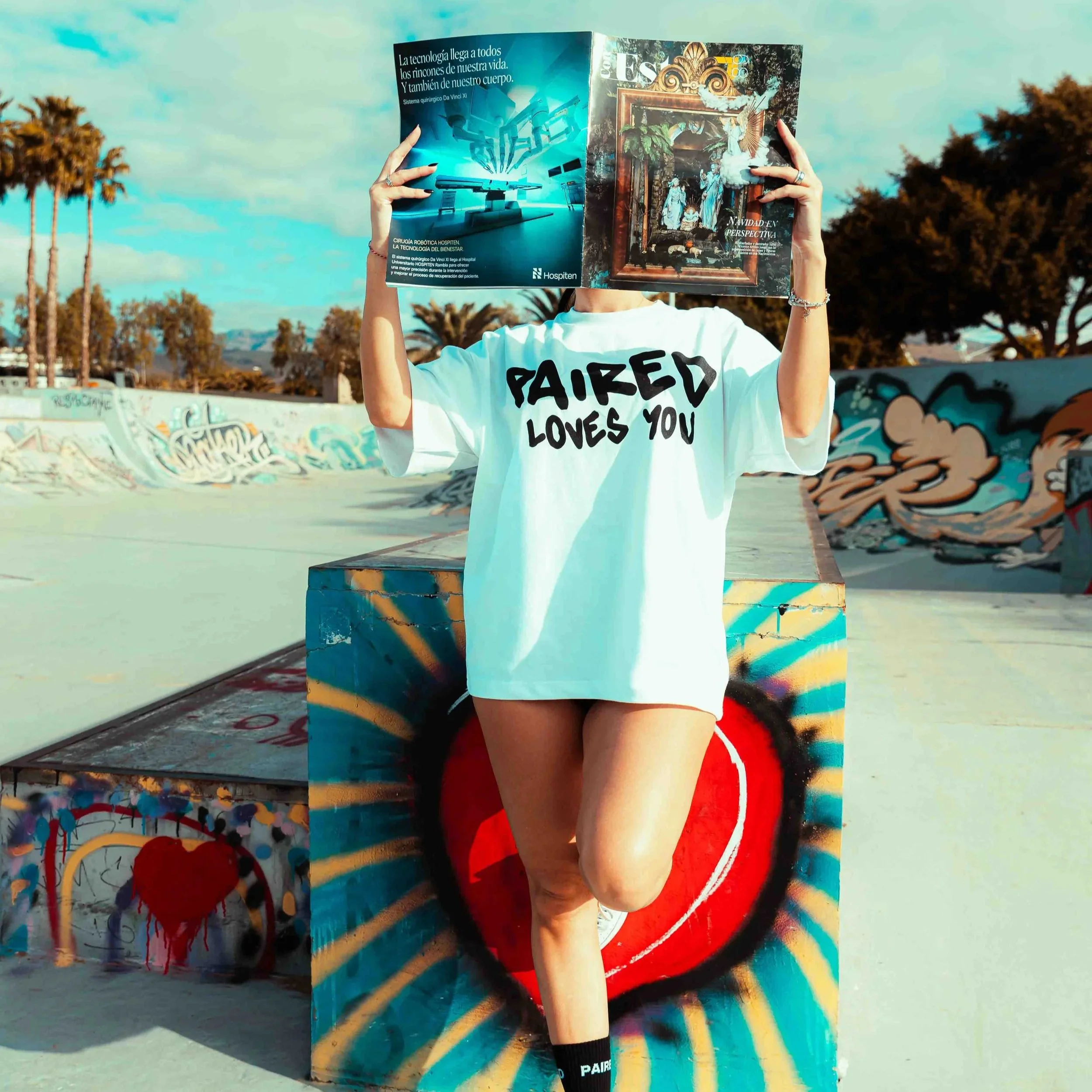 Person holding an open magazine in front of their face at an outdoor skate park with graffiti, trees, and a blue sky in the background. The person is wearing a white t-shirt with black lettering and red shorts.