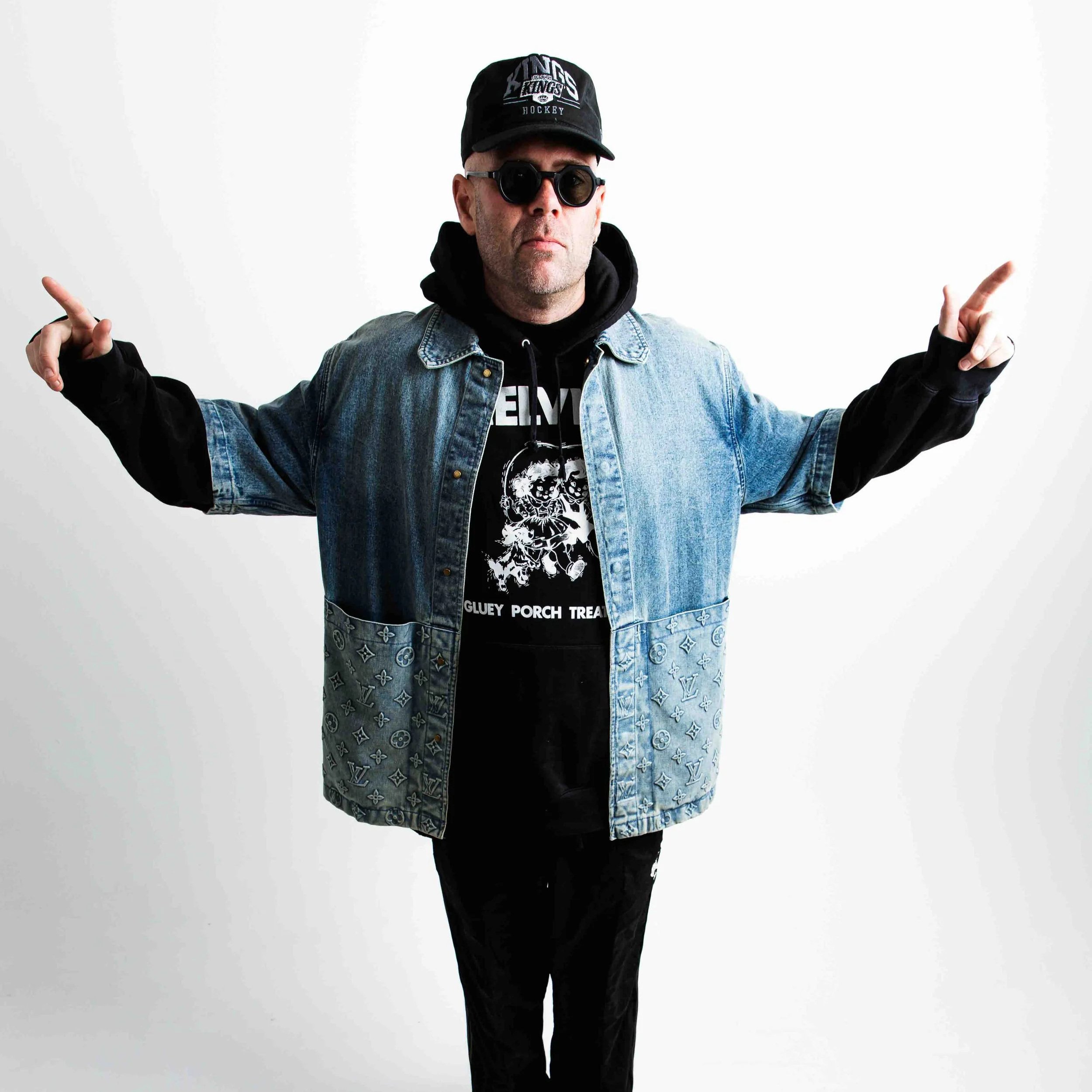Solardo wearing a black cap with sports logo, black sunglasses, a black hoodie, a denim jacket with designer print, and black pants, standing against a white background.