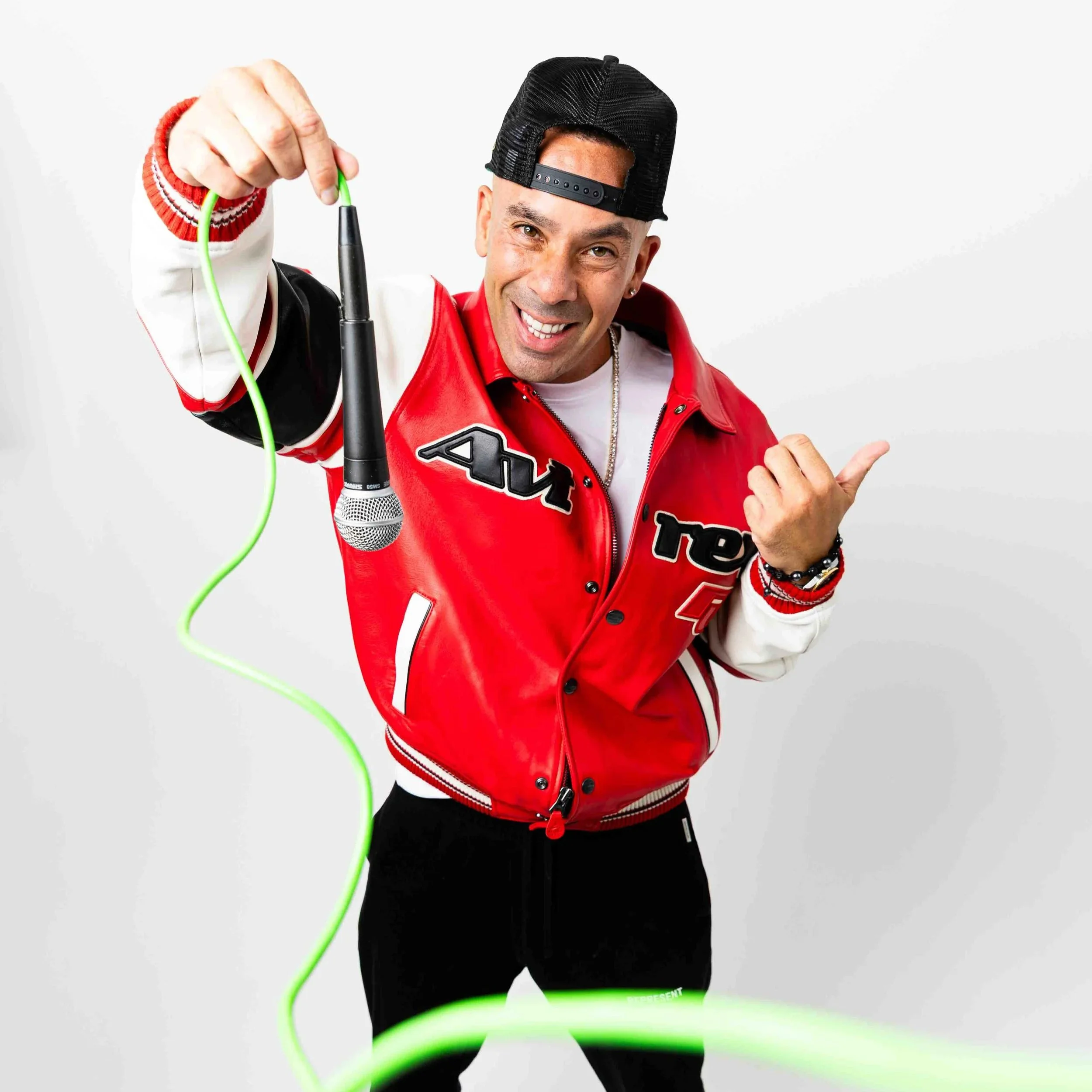 Solardo with light skin wearing a black trucker hat turned backwards, a red and white varsity jacket, and a white T-shirt. He is smiling and posing with a finger pointing outward. The background is plain white, and there is a green cord or wire cross