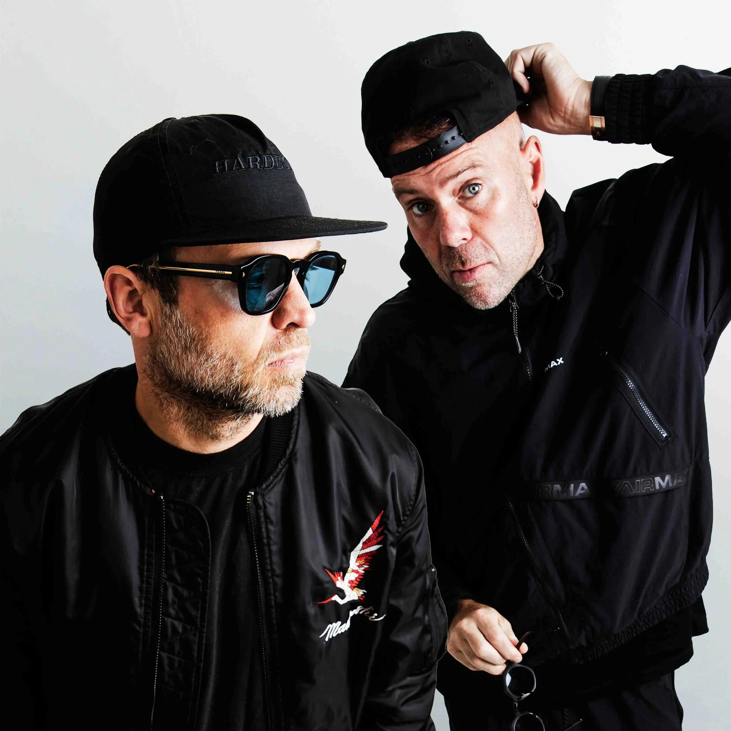 Solardo wearing black jackets and black caps, one with sunglasses, are close to each other against a plain light gray background. One man is touching his cap, the other is holding sunglasses.