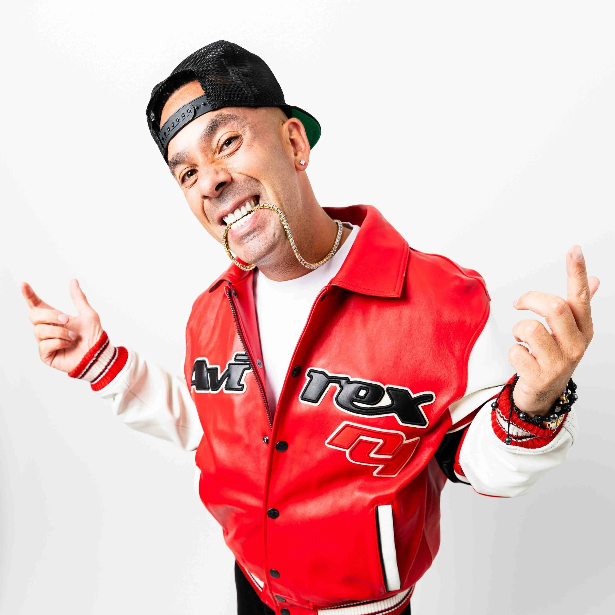 A young man wearing a black baseball cap backward, a red and white varsity jacket, and jewelry, smiling and making peace signs with both hands, against a plain white background.