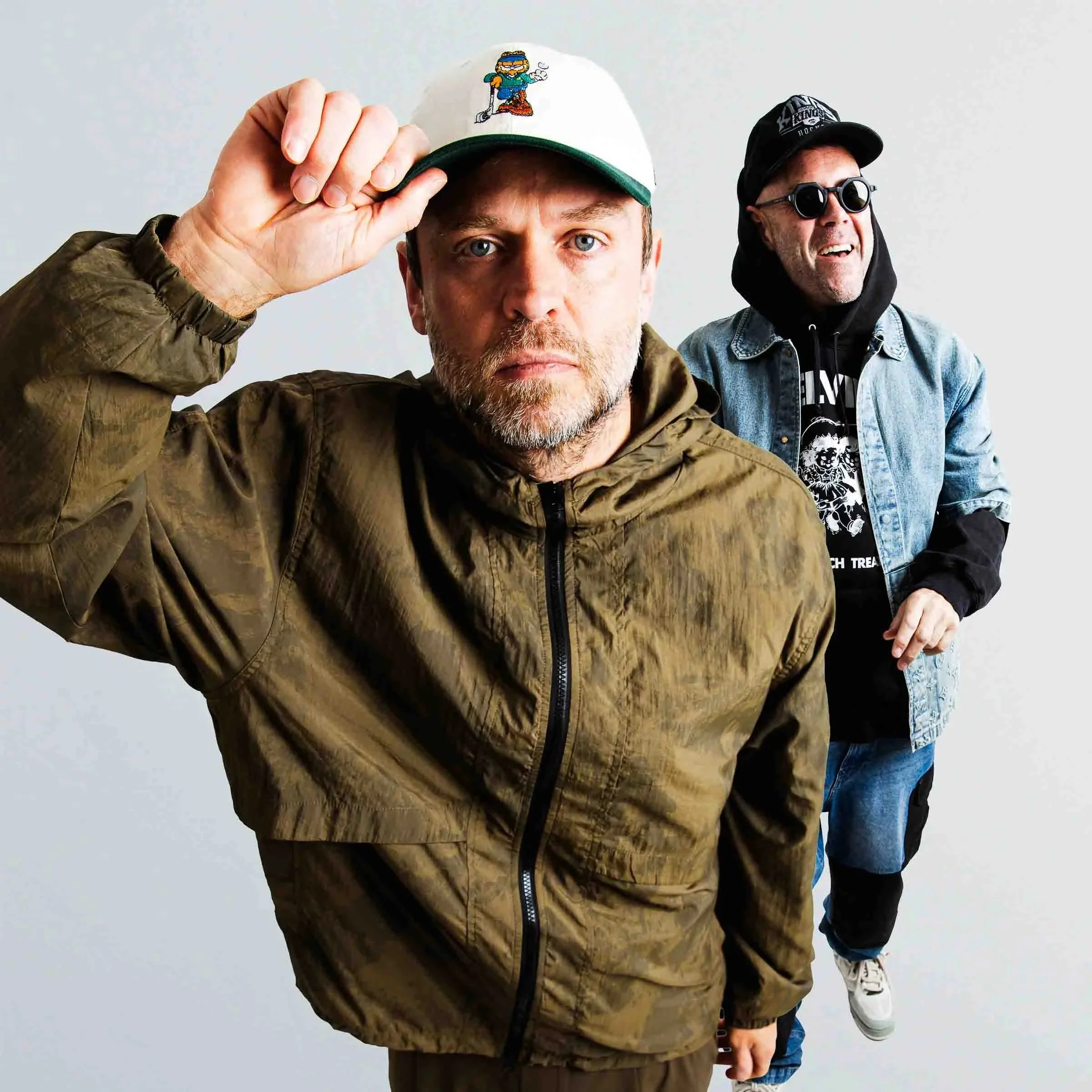 Solardo standing side by side, with one man in the foreground adjusting his white baseball cap featuring a cartoon character, and the other man in the background wearing sunglasses and a baseball cap. The background is plain and neutral.