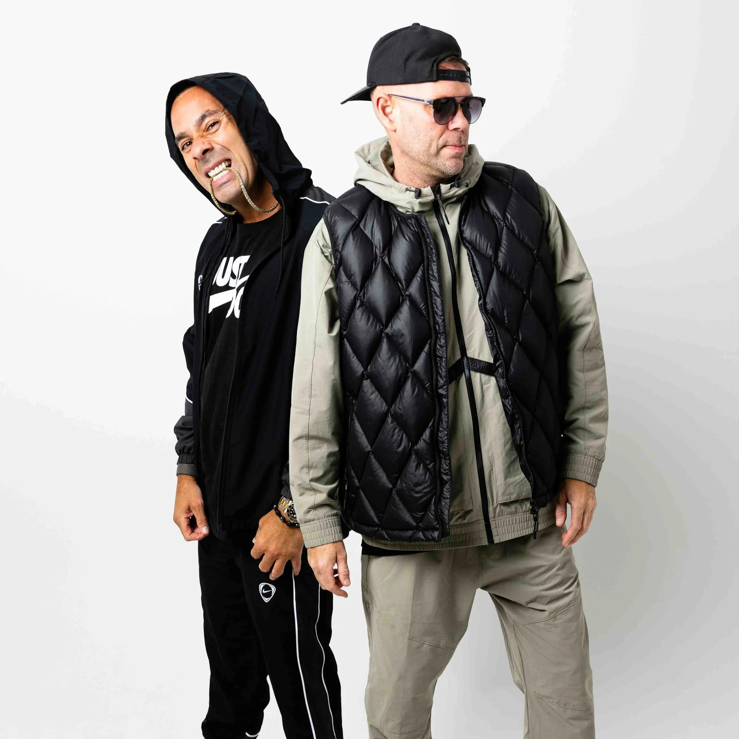 Solardo standing side by side, one with a hooded jacket showing a gold chain and smiling with a gold grill, the other with sunglasses, a cap worn backward, and a puffer vest, against a plain white background.