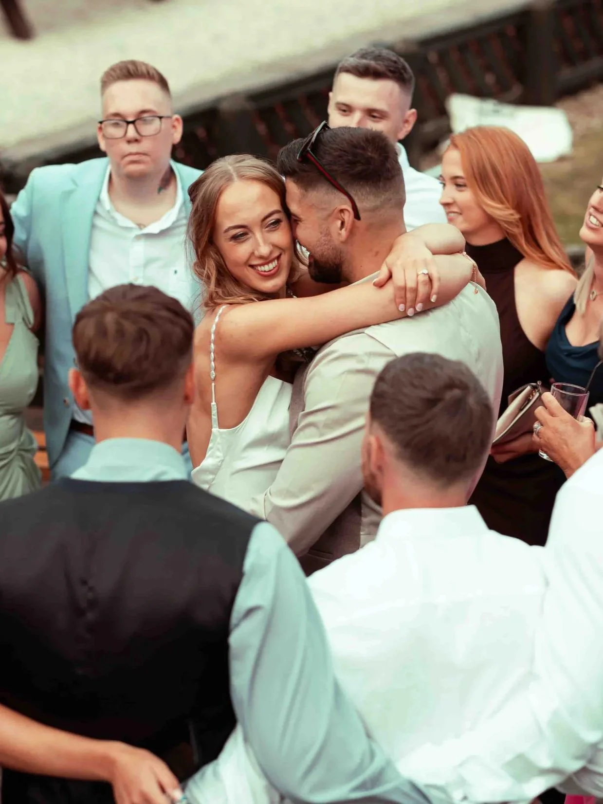 Couple sharing an intimate moment, hugging and smiling, while celebrating at a wedding reception with family and friends around them.