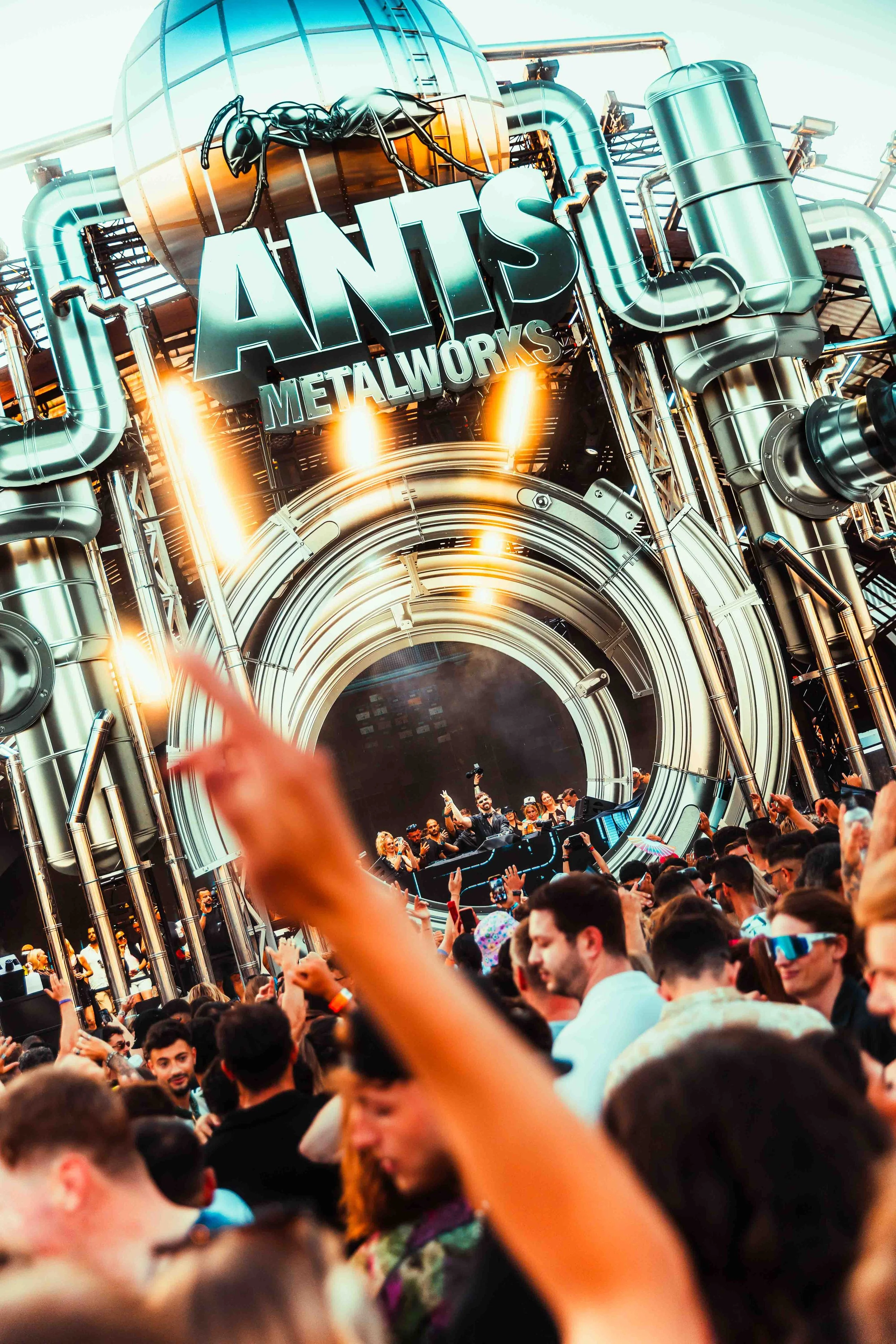 Crowd of people at the Acceptance of Art Festival at an outdoor event with a metallic stage featuring large pipes, lights, and a sign that reads 'ANTS METALWORKS' with a giant ant sculpture on top.