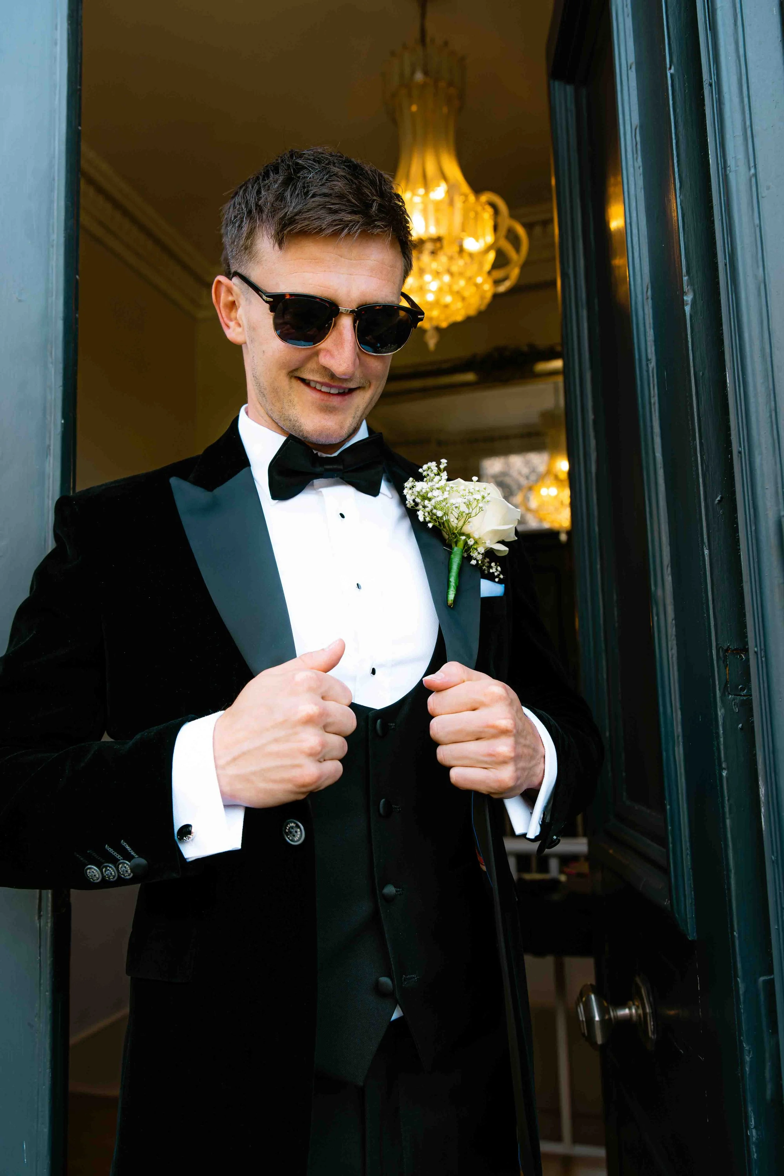 A man in a tuxedo with sunglasses and a boutonniere opening a black door, smiling.