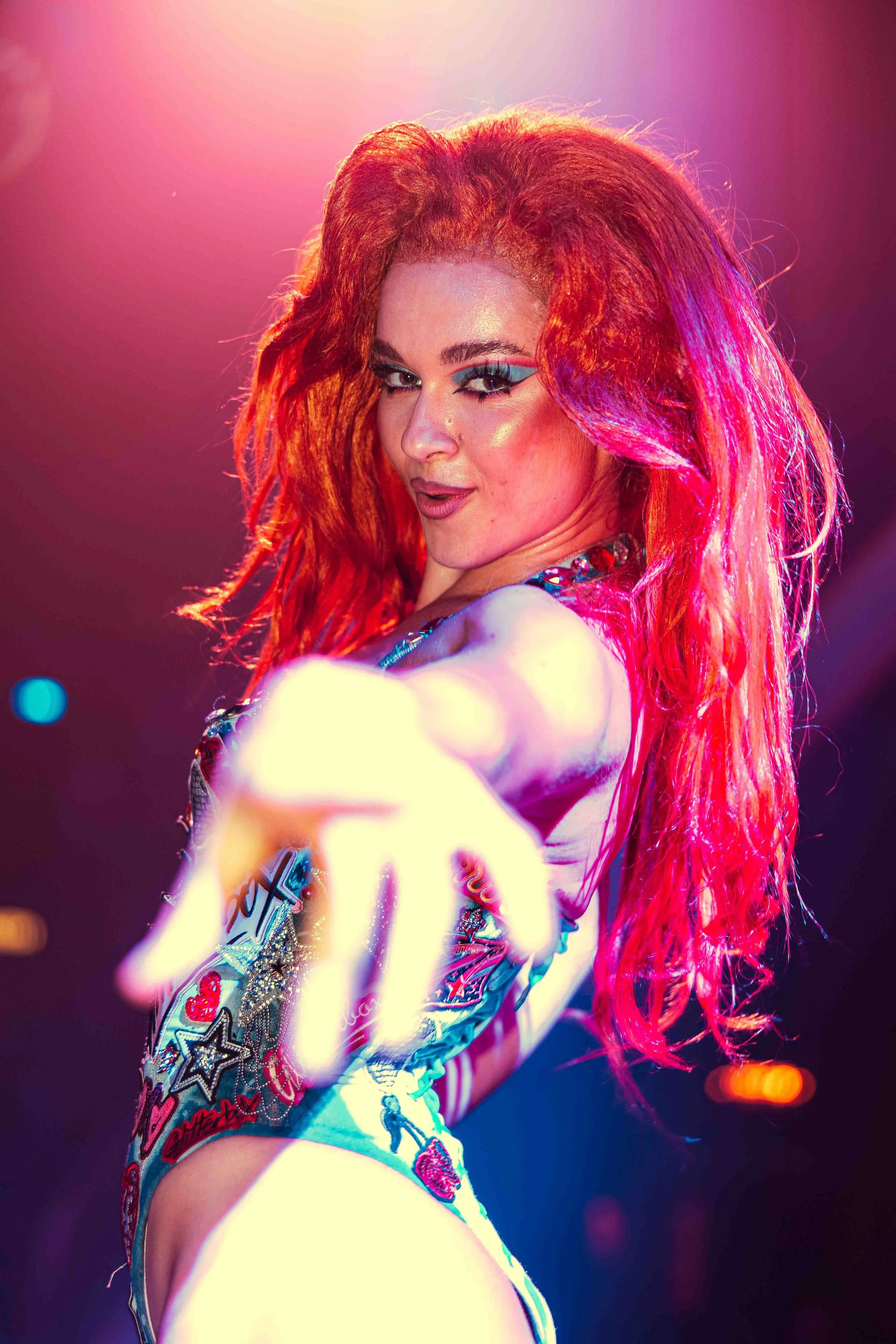 A woman with vibrant red hair styled in loose waves, wearing bold makeup with turquoise eye shadow, posing confidently with a slight smile on stage with colorful lighting.