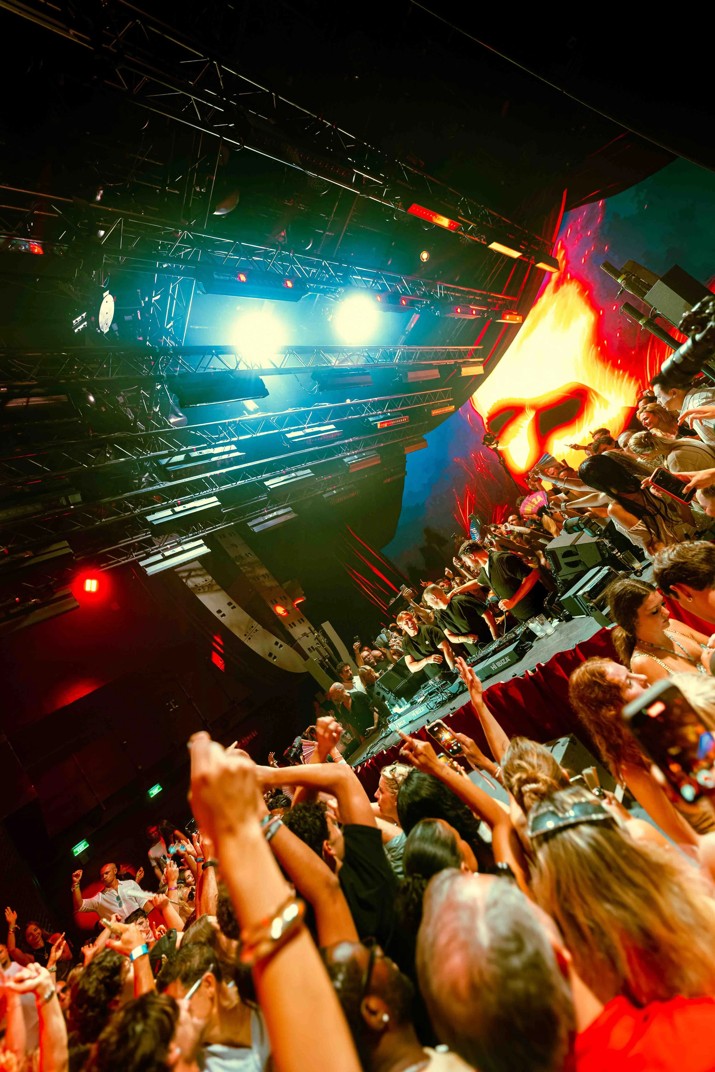 Crowd of people at a concert or festival with stage lights and large digital screen displaying a fiery volcano or lava eruption.