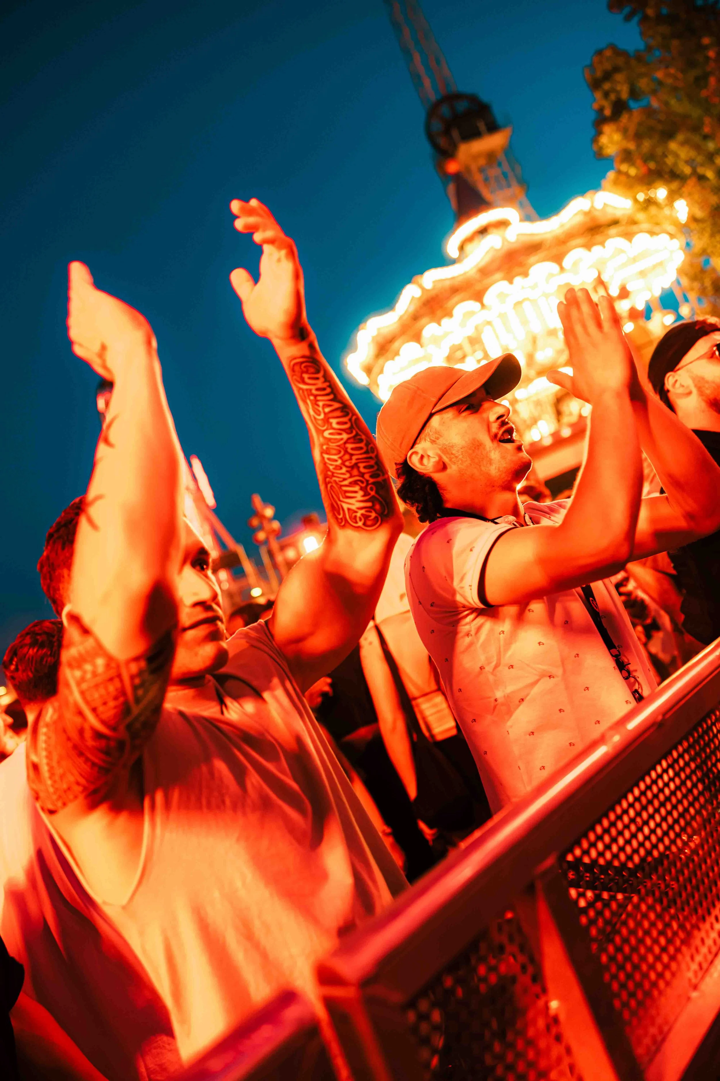 People at an amusement park enjoying a nighttime ride, with amusement park rides and lights in the background, and their hands raised in the air.