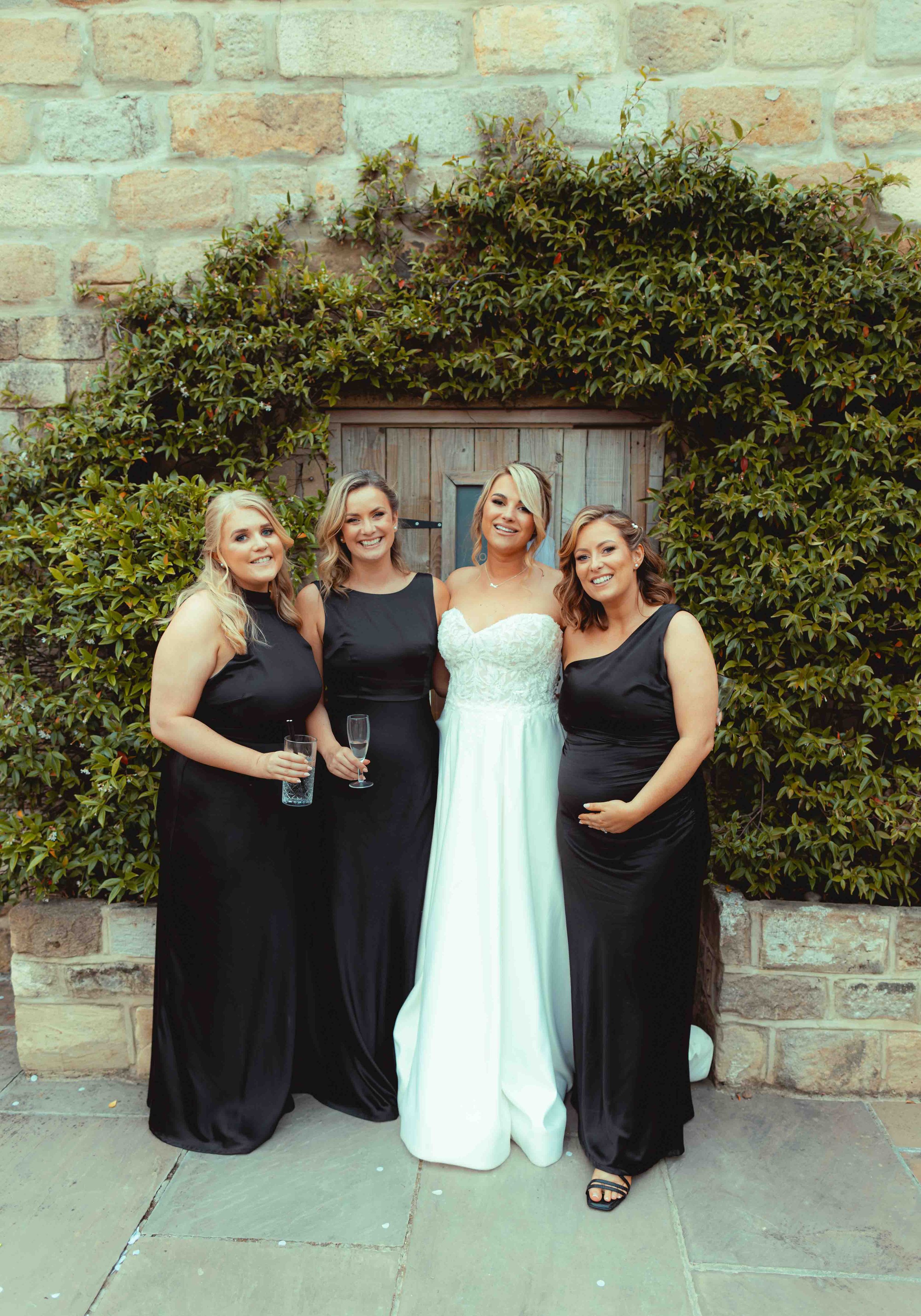 Four women standing together outdoors in front of a brick wall and a wooden door, smiling at the camera. The woman second from the right is wearing a white dress, and the other three women are wearing black dresses. Two women are holding champagne gl