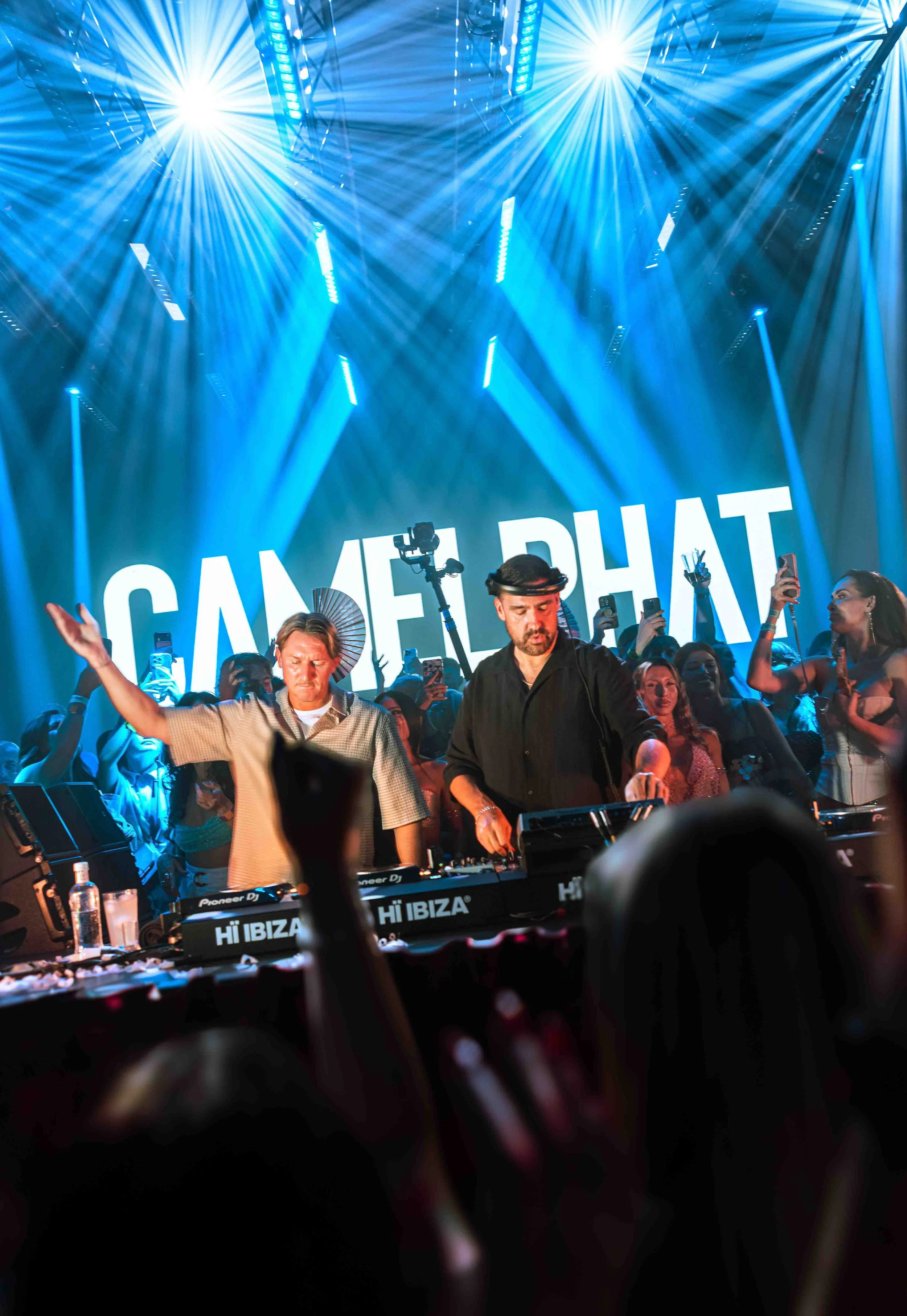 A DJ duo performing at a lively event with bright blue stage lighting and a large crowd. The background features a sign that reads 'GAME PLAN.' The crowd is engaged, with some holding phones and others dancing.