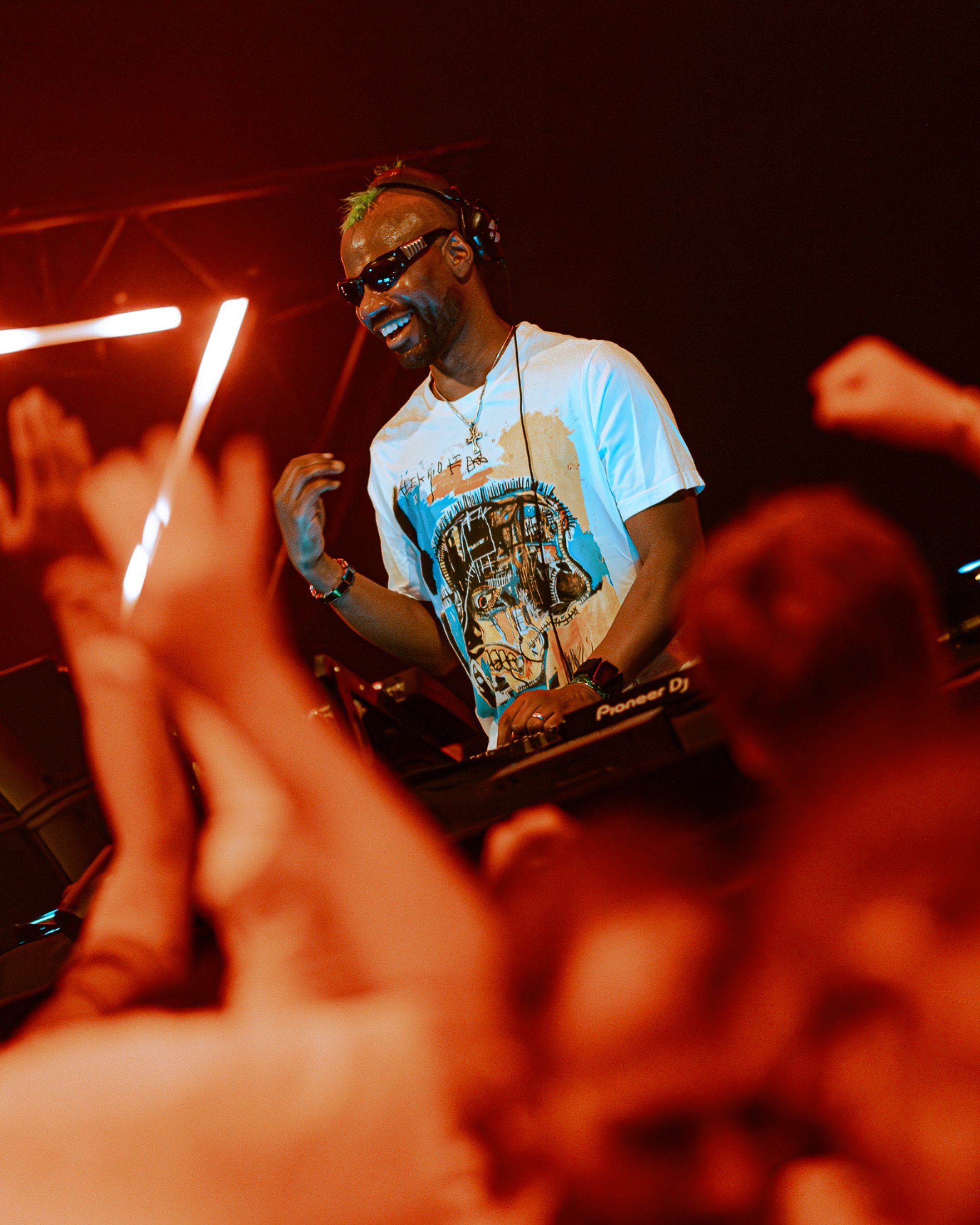 Green velvet, wearing sunglasses and a graphic T-shirt, performing at a party or concert with red and orange lighting. The DJ is smiling and using DJ equipment labeled 'Pioneer DJ'.
