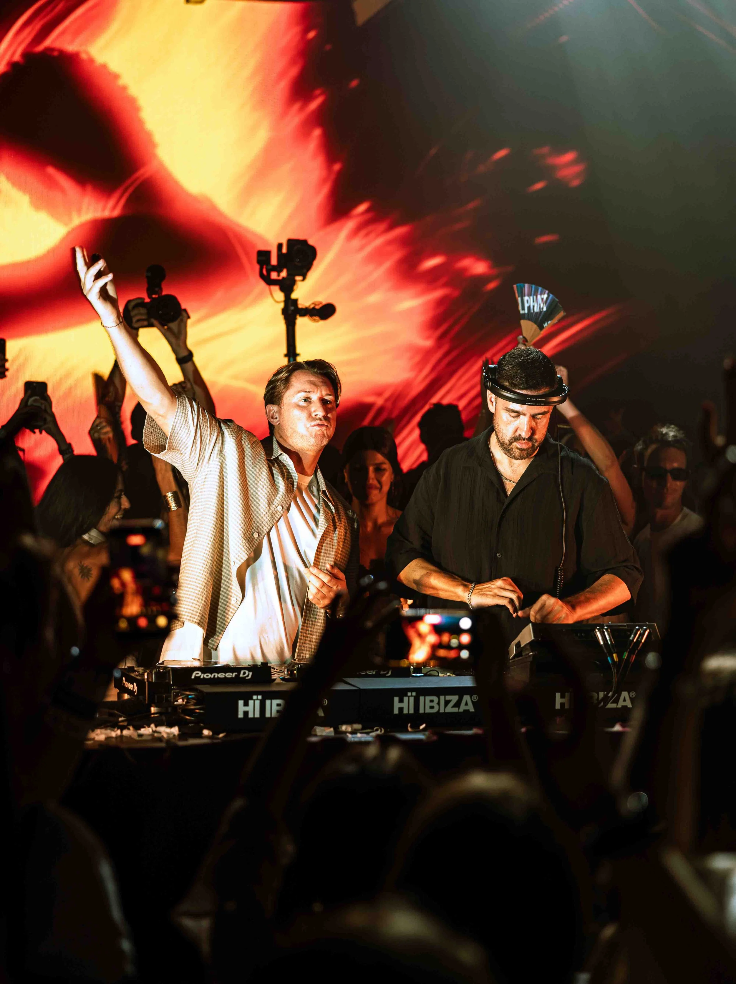 A DJ with a headset and a man with his arm raised dancing at a party with a vibrant, colorful digital backdrop.