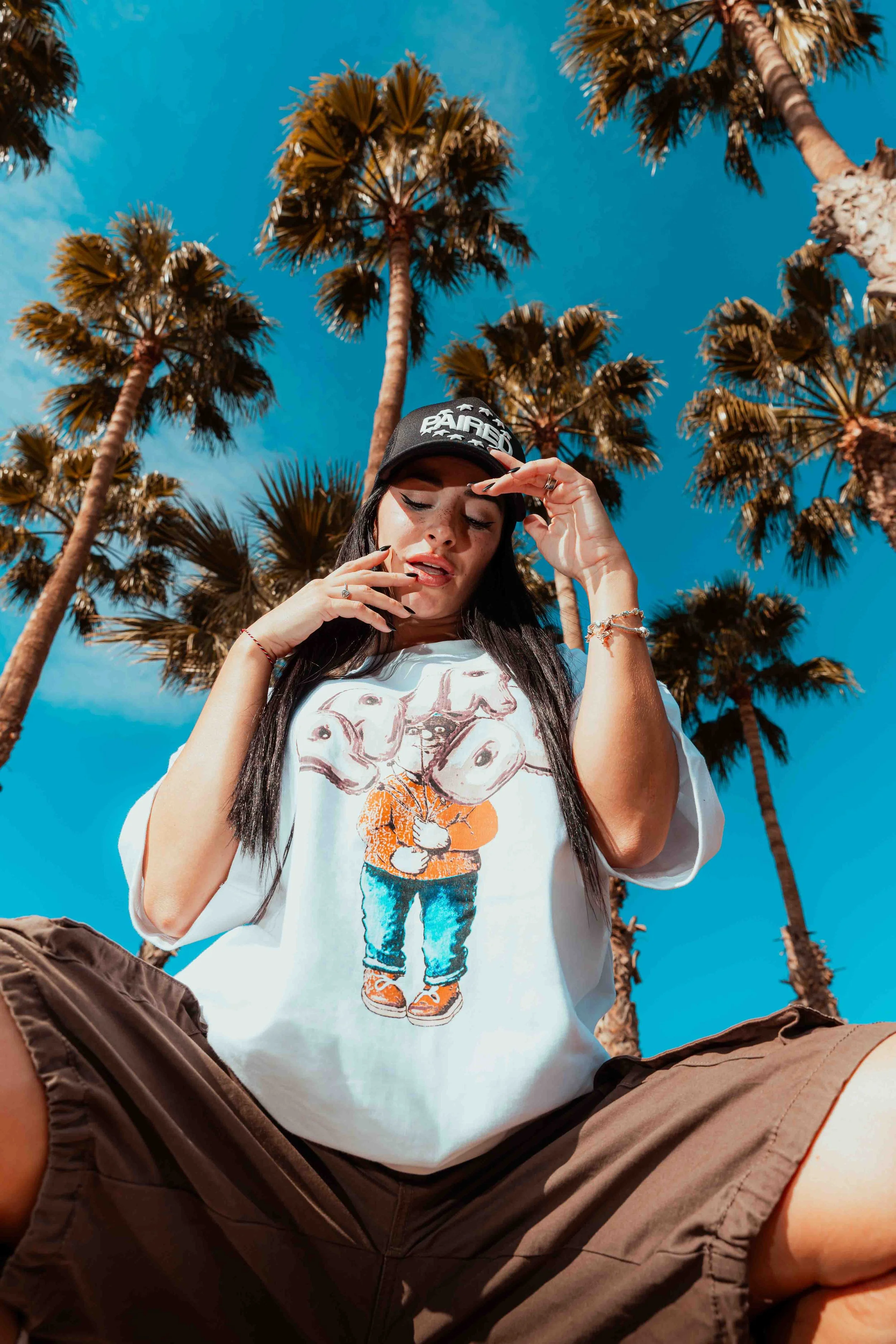 Young woman sitting outdoors with palm trees in the background, wearing a baseball cap and a graphic t-shirt, looking down and touching her face.
