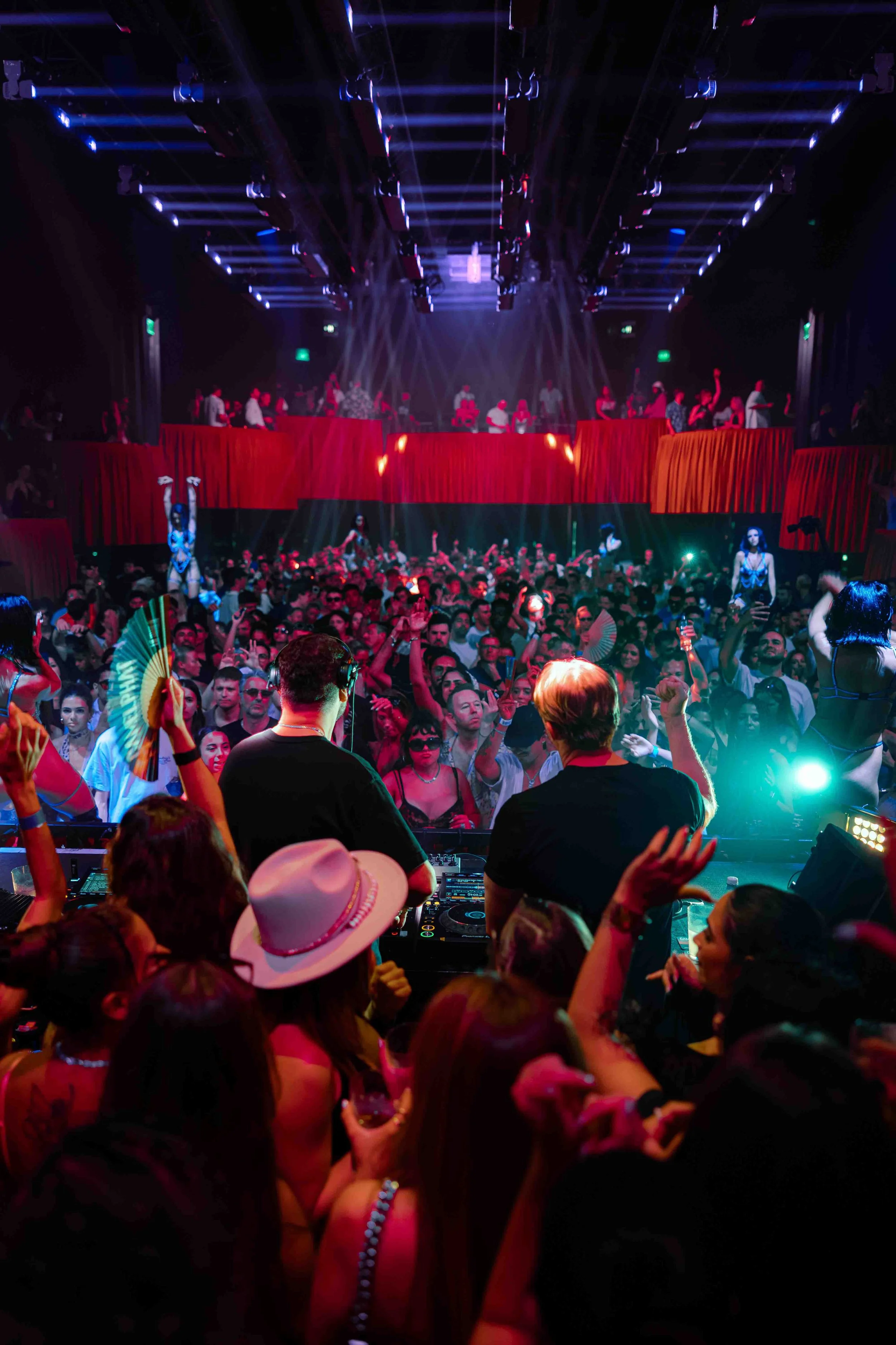 A crowded dance club with a DJs Camelphat at the turntables in the foreground, colorful lighting, people dancing with raised hands, and a raised stage with performers and red curtains in the background.