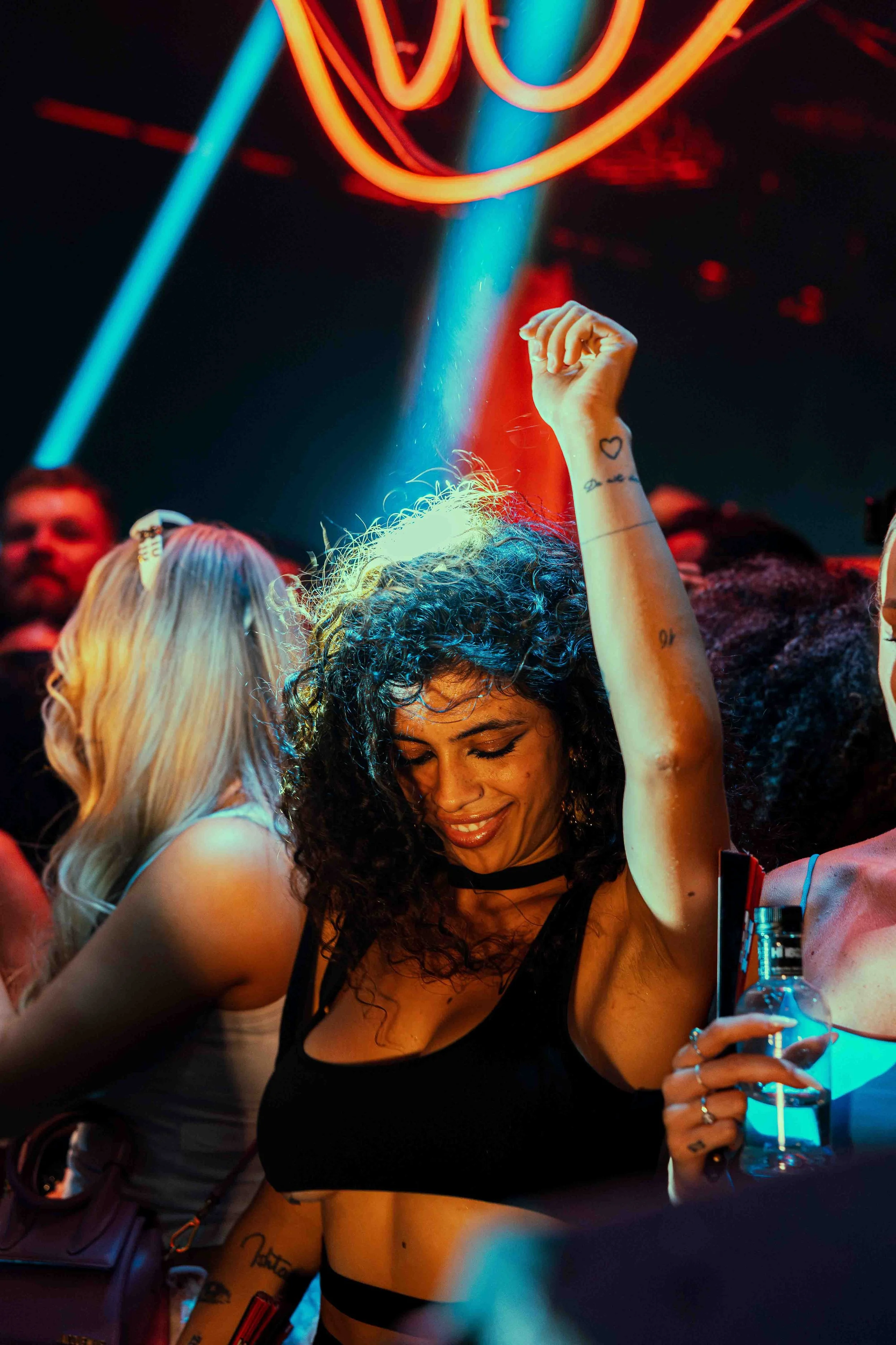 A woman with curly dark hair dancing in a club, holding a drink, with colorful neon lights overhead and other people around her.