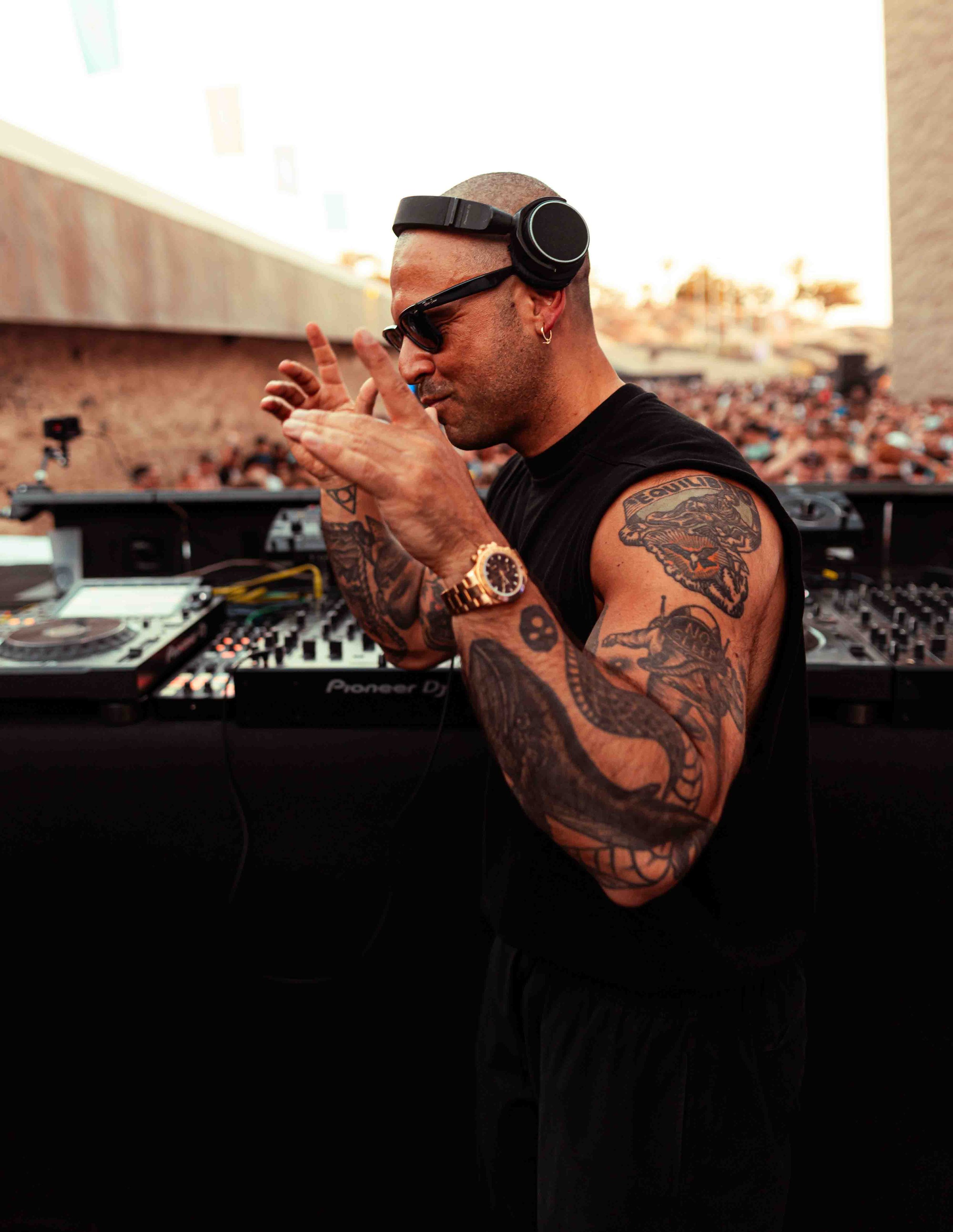 A tattooed male DJ with a shaved head, wearing sunglasses and headphones, playing music.