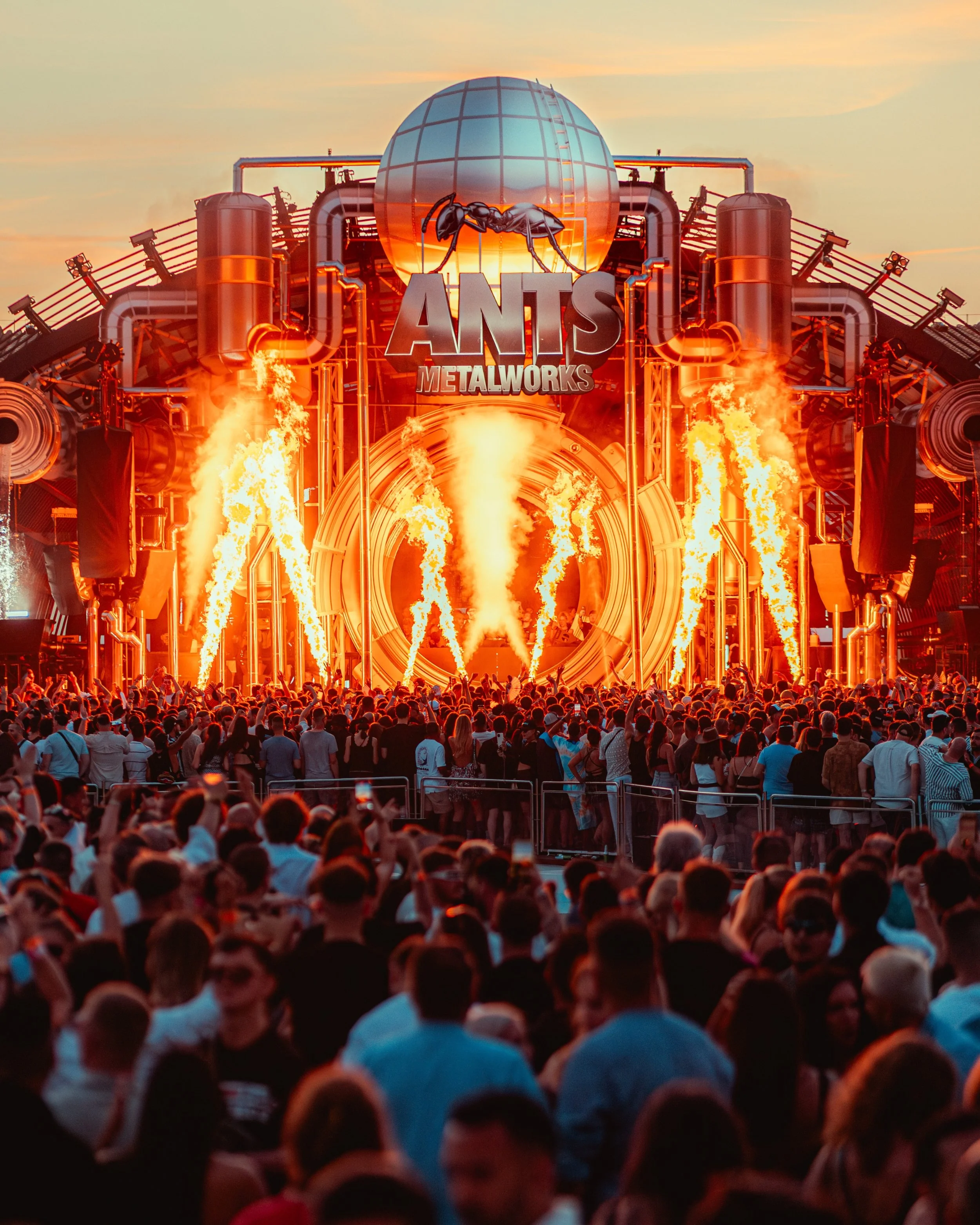 A large crowd watching a live music or DJ performance at an outdoor event in the evening. The stage features the Ants Metalworks logo, with flames and pyrotechnics. The sky is partly cloudy with a sunset glow during Patrick Toppings CLosing set