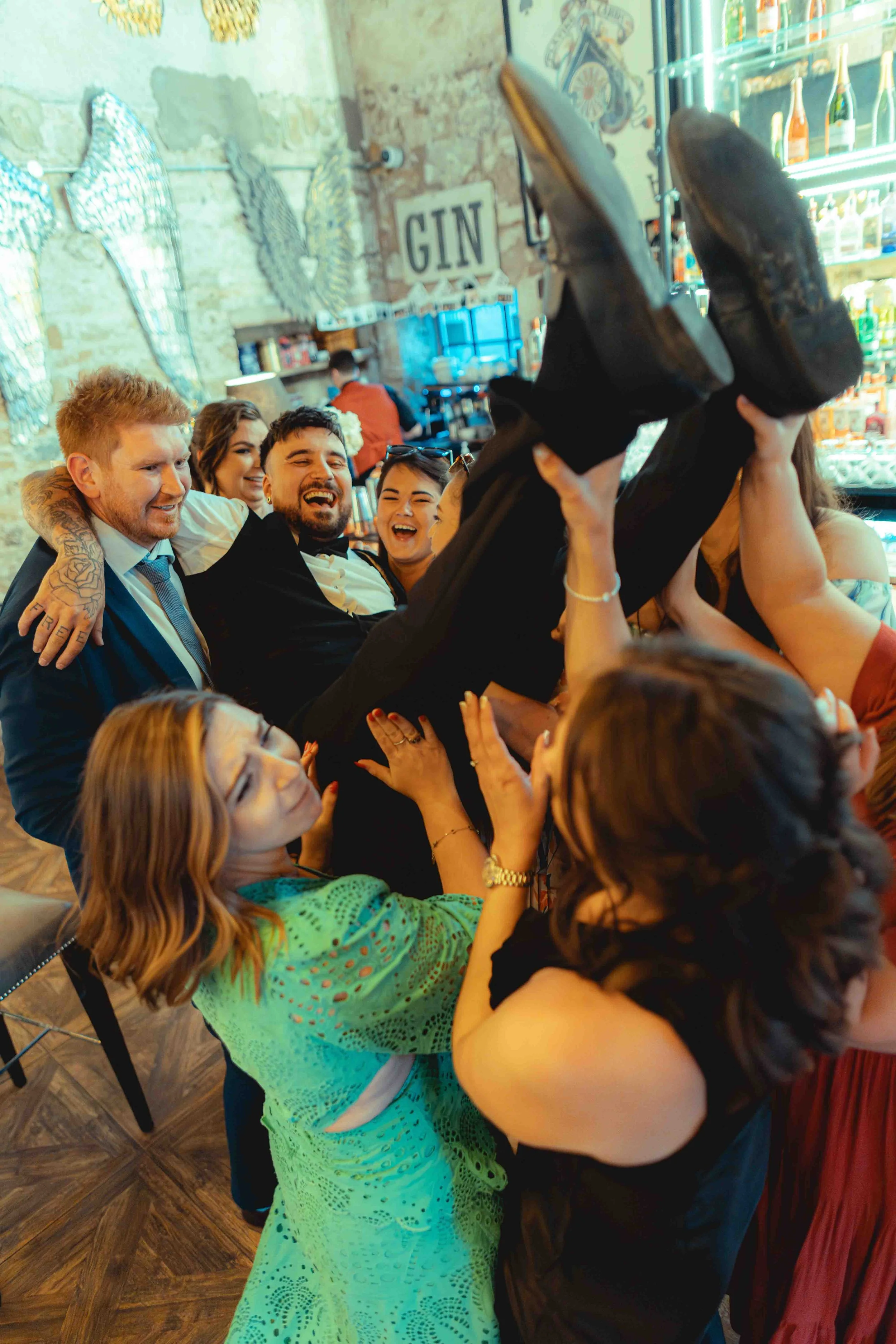 Group of people laughing and celebrating, with some individuals lifting others in the air, in a lively indoor setting decorated with wings and a sign that says 'GIN.'