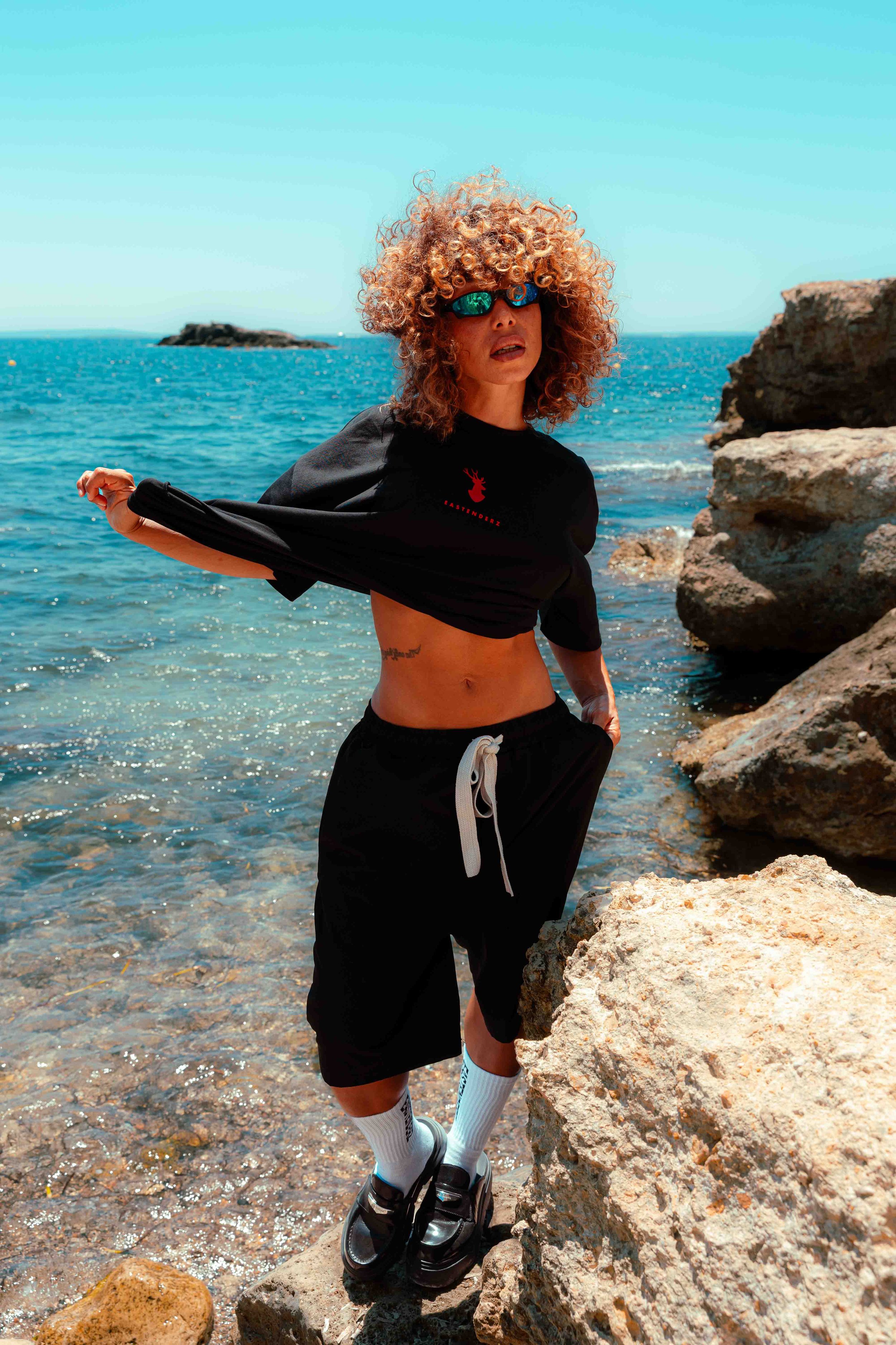 A person with curly blond hair wearing sunglasses, a black t-shirt with a red deer logo, black shorts, white socks, and black sneakers standing on rocky shoreline by the sea, with rocks and water in the background.