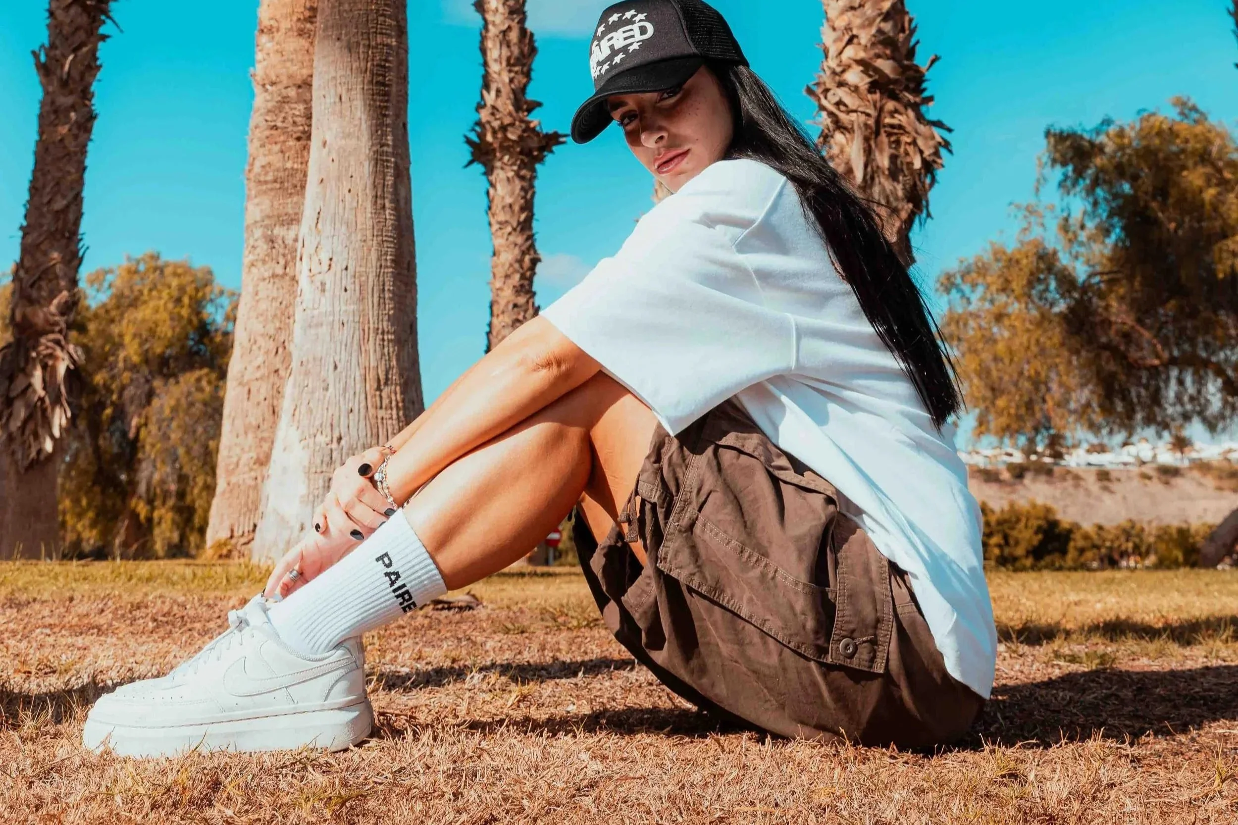 Young woman sitting outdoors in front of palm trees, wearing a black cap, white t-shirt, brown shorts, white sneakers, and white socks, with long black hair and looking at the camera.