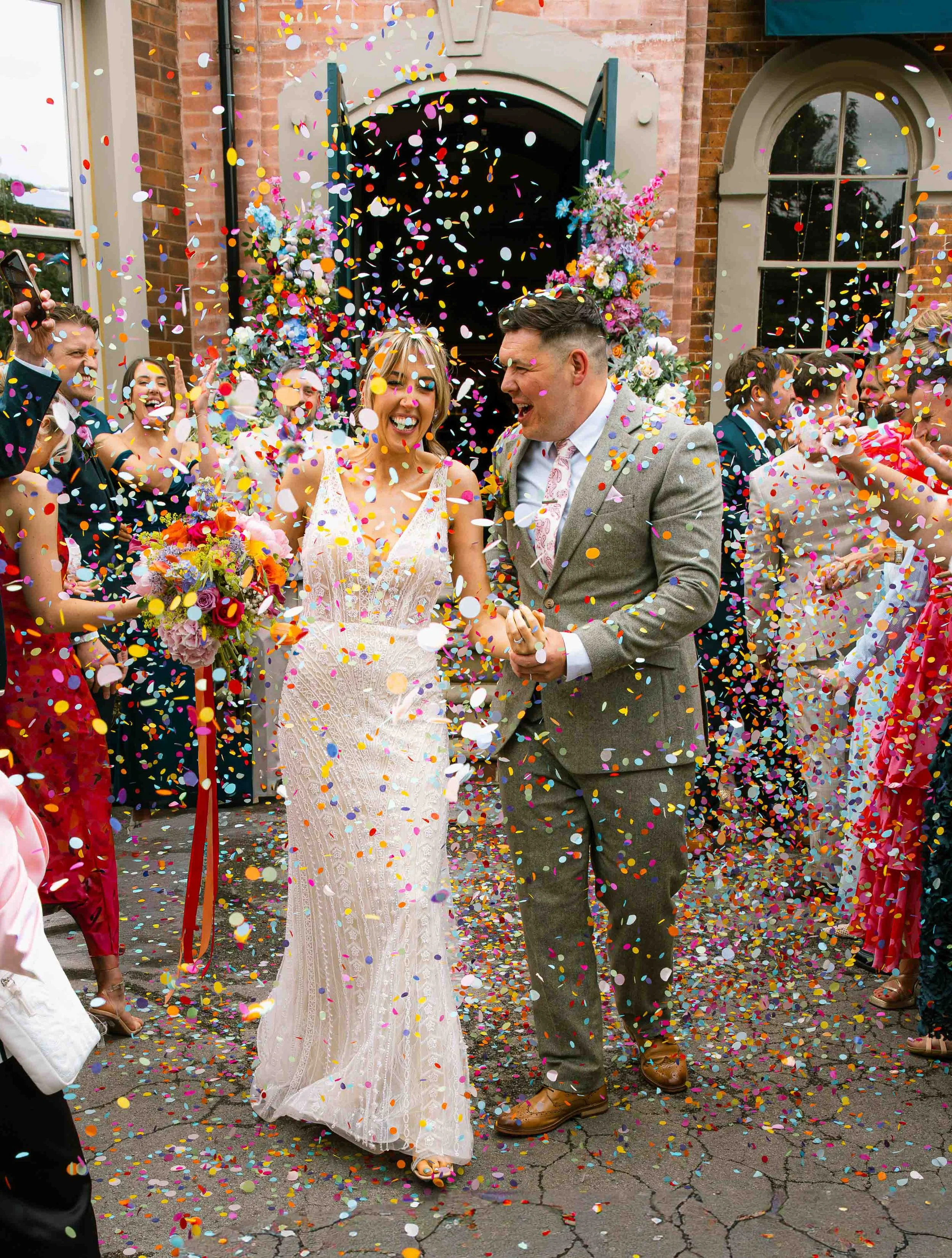 A bride and groom celebrating their wedding outdoors surrounded by friends and family, with colorful confetti falling around them. The bride is wearing a white wedding dress and holding a bouquet of flowers, and the groom is dressed in a gray suit. T