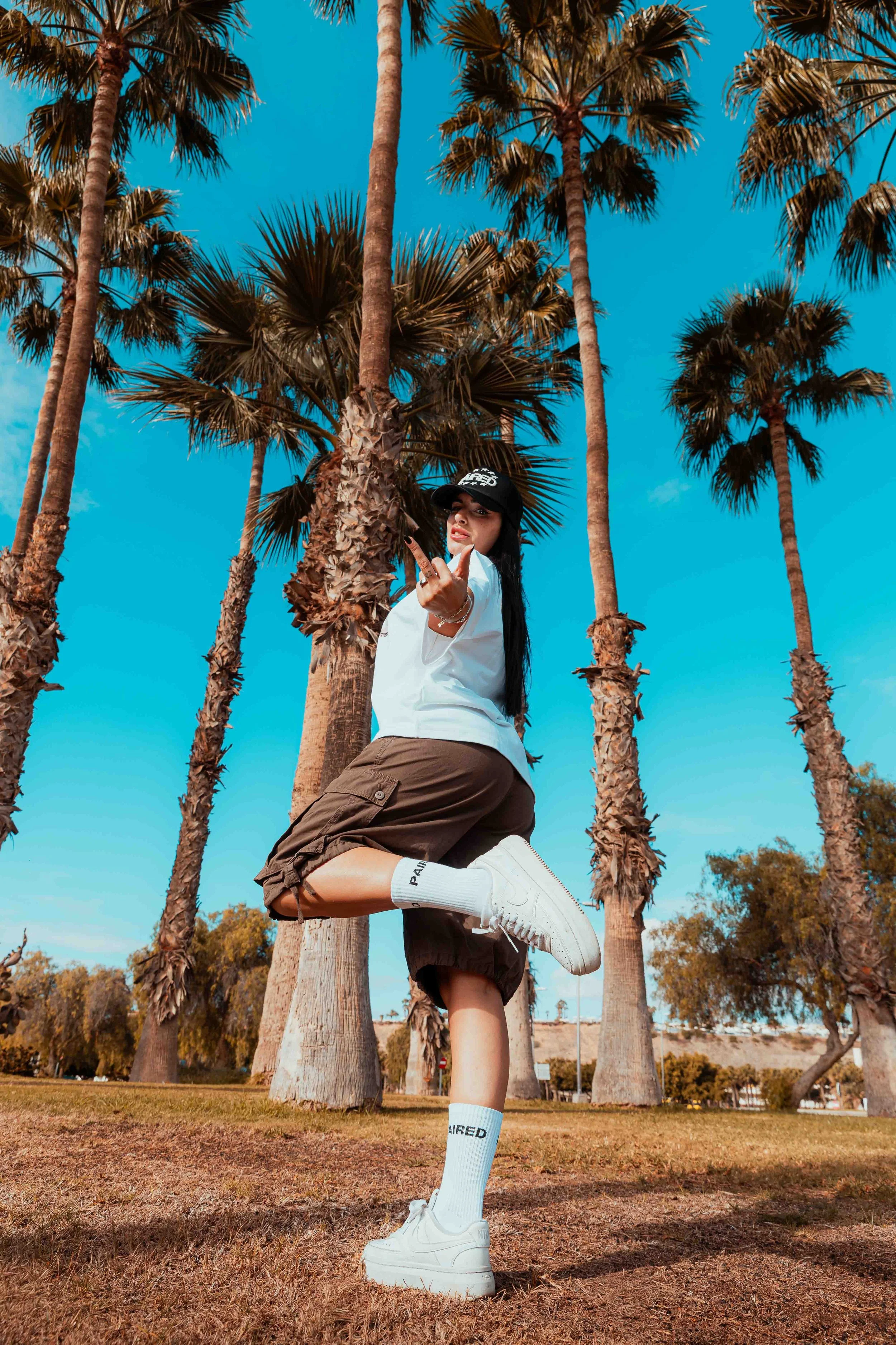 A woman standing on one leg among tall palm trees outdoors, wearing white sneakers, white crew socks, brown shorts, a white T-shirt, a black baseball cap, and sporting dark hair.
